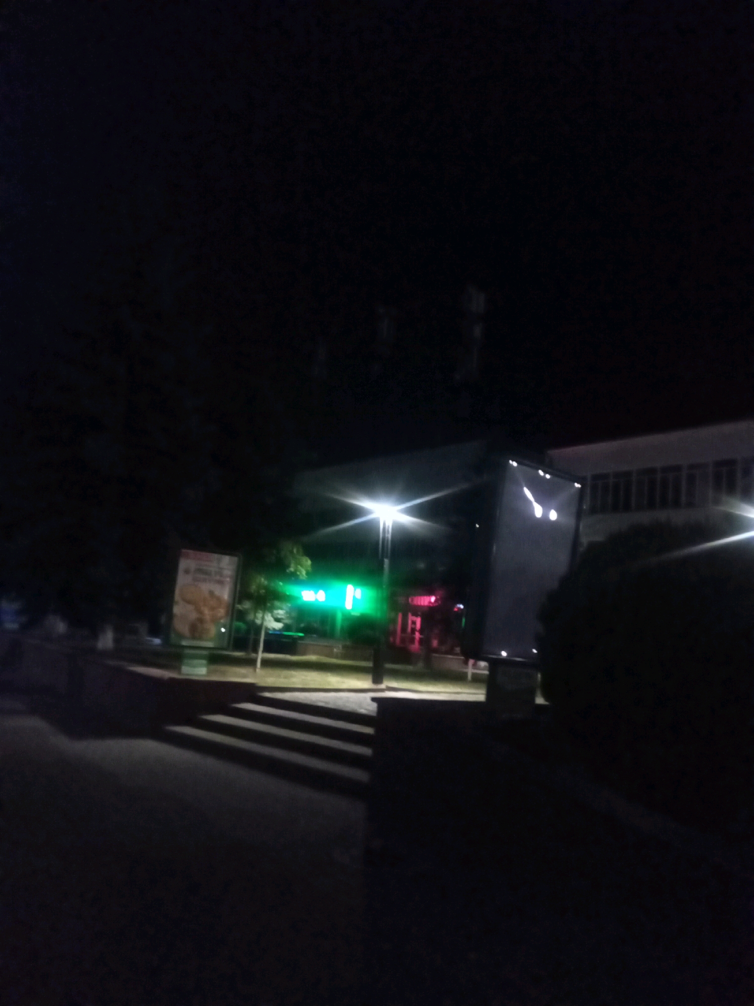 The image shows a dark outdoor scene at night, possibly in Tiachiv, Ukraine, with artificial lighting creating an eerie atmosphere. In the center of the frame, a large, illuminated digital billboard stands angled towards the viewer, displaying white symbols that resemble a face or abstract shapes. To the left of the billboard, a lighter-colored billboard is partially visible, with text and an image, likely an advertisement. Behind these, a building with multiple windows is faintly discernible against the dark sky. Green neon lights emanate from a structure to the left, possibly a business or establishment. A streetlamp with a bright light is visible to the right of the digital billboard, casting long shadows. In the foreground, a dark, paved area, likely a street or sidewalk, is present, with a set of concrete stairs leading up to a raised area. Trees and foliage are silhouetted against the light sources, contributing to the shadowy ambiance. The overall impression is that of a commercial or public area after closing hours, illuminated by artificial lights.