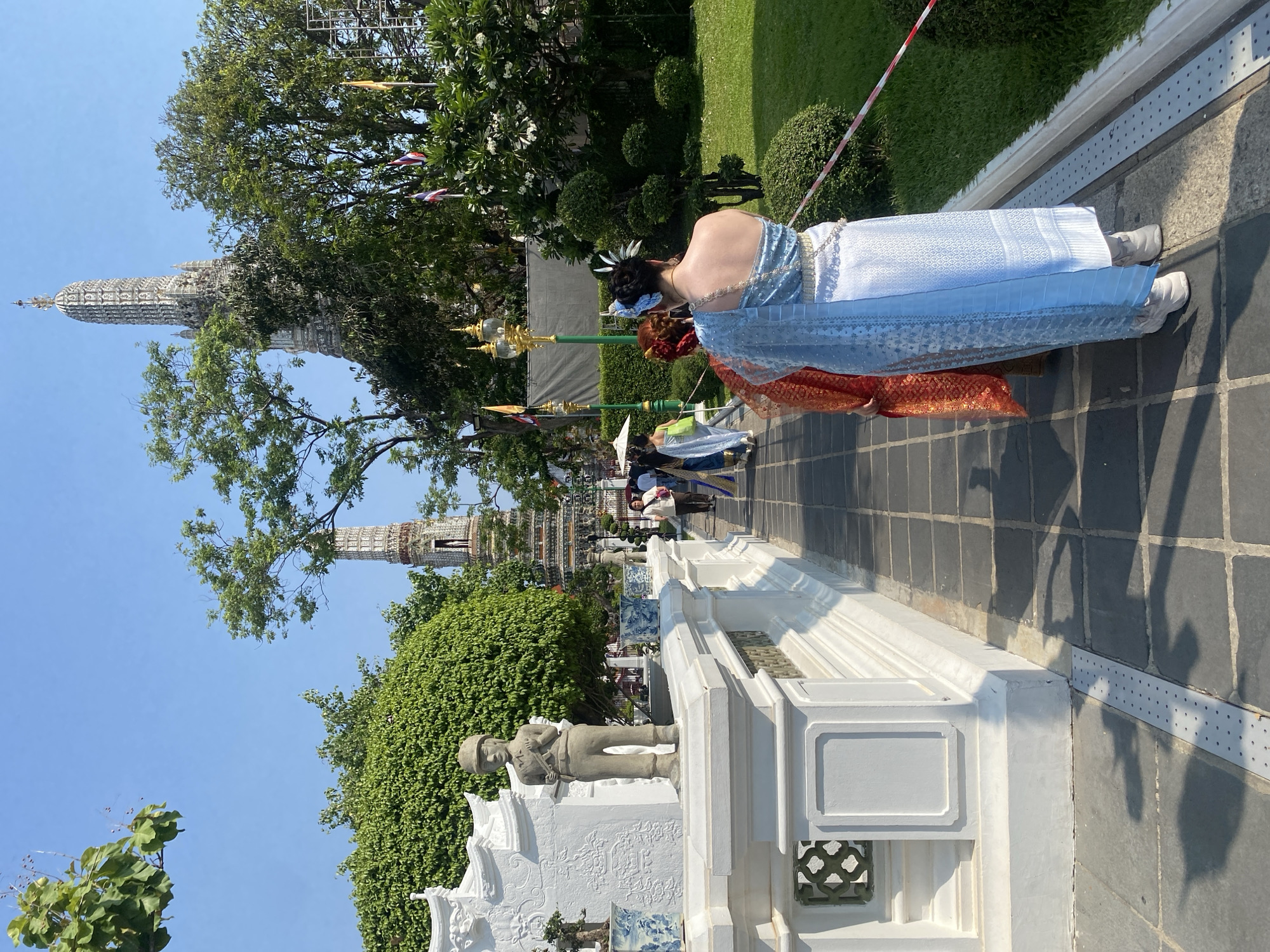 A daytime shot depicts a woman in a traditional Thai dress walking along a pathway, with the Wat Arun temple visible in the background, suggesting the location is in Bangkok, Thailand. The woman's dress features a light blue fabric, and she is accompanied by another person in a similar traditional garment. The pathway is paved with dark grey tiles, and a white wall runs alongside it, decorated with sculptures and intricate designs. Lush green trees and bushes surround the path, contributing to the outdoor ambiance. The sky is clear and blue. Several people, including tourists, are further down the pathway.