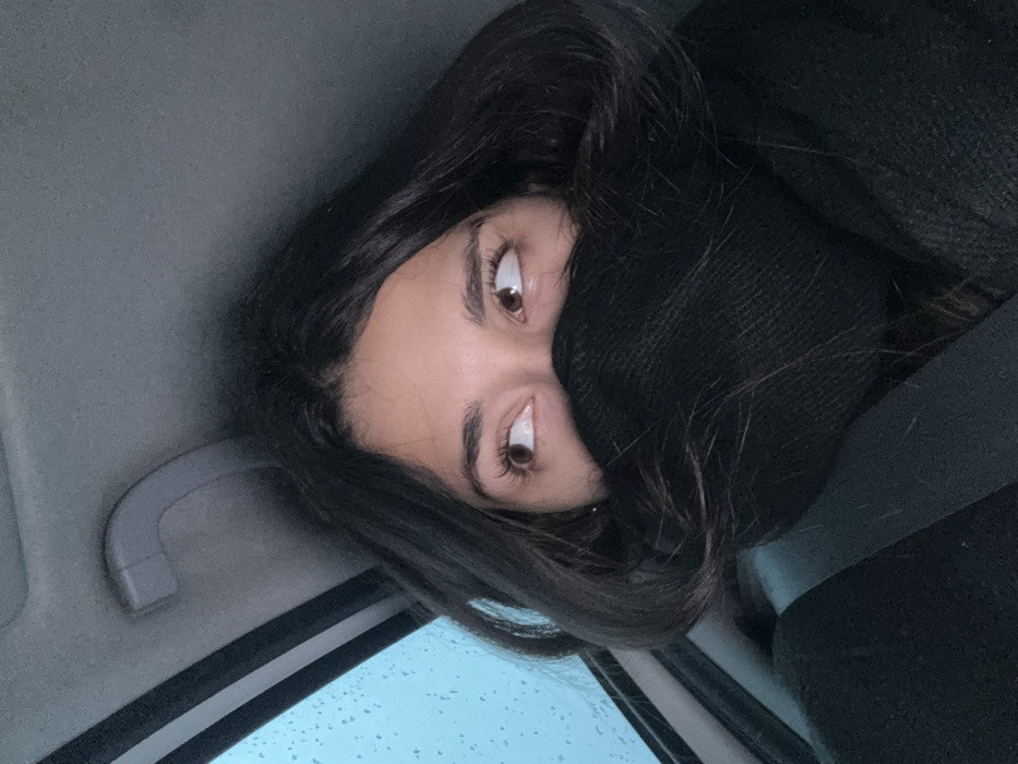 The scene depicts a female individual occupying a passenger seat within a vehicle, situated in Cibukovac, Serbia. The woman's face is partially obscured by a dark knitted garment, likely a scarf, covering her nose and mouth. Her brown eyes are visible, gazing upwards and to the viewer's left. Dark hair frames her face. A black seatbelt is visible diagonally across her left shoulder and chest. The vehicle's interior features a grey headliner and a grey overhead grab handle. Through a window positioned on the left side of the frame, raindrops are discernible against an ambient light-blue background, indicating wet weather conditions.