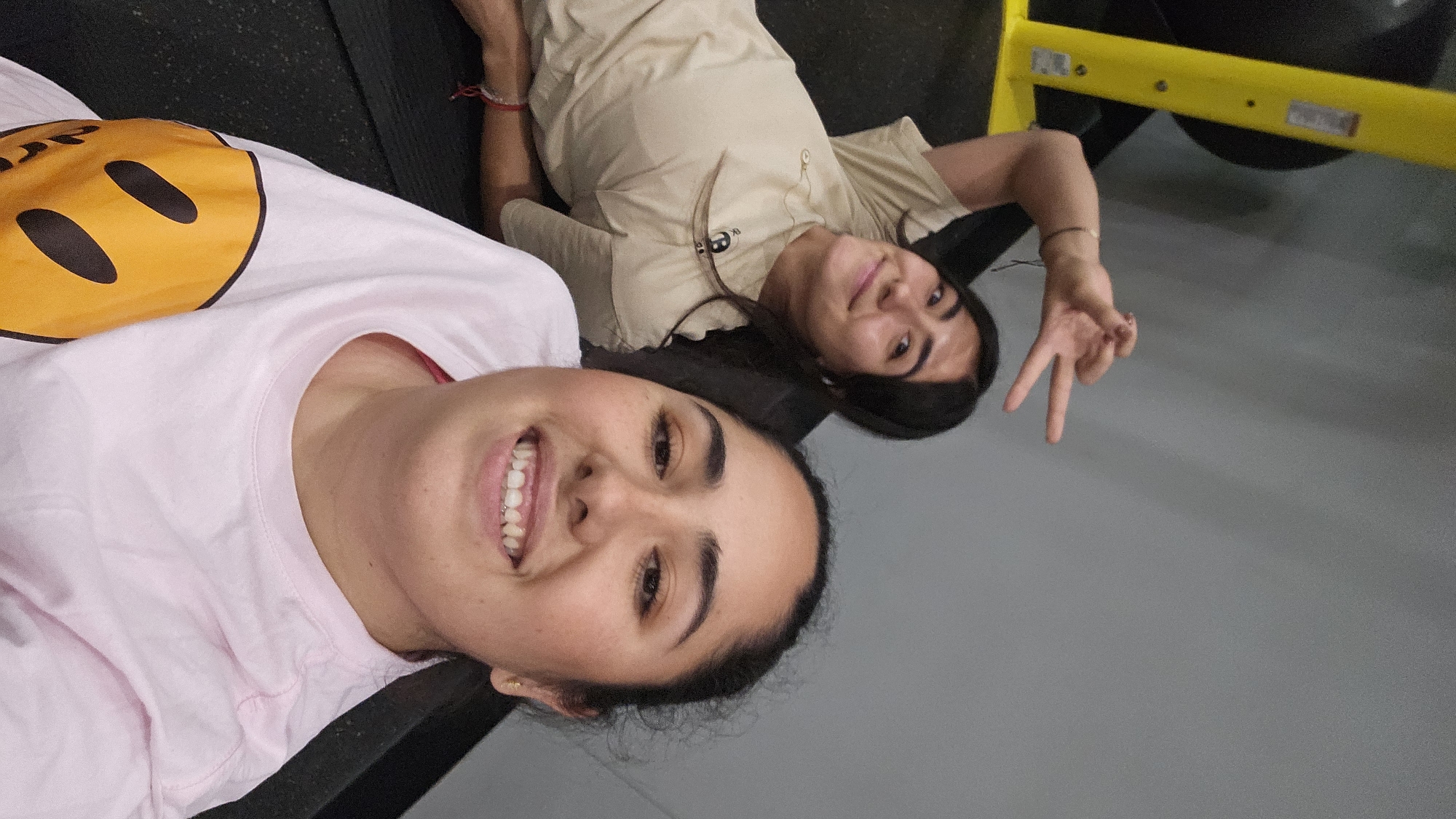The image displays two young women lying down on what appears to be gym equipment, looking upwards towards the camera, likely taking a selfie in a gym located in Puebla, Mexico.
In the foreground, one woman is wearing a light pink short-sleeved t-shirt adorned with a large yellow smiley face logo. The word "drew" is partially visible within the smiley, indicating it is a Drew House brand item. She has dark hair, dark eyebrows, and a wide, cheerful smile, revealing dental braces on her upper teeth. A small gold earring is visible on her left earlobe.
Behind her, the second woman is also smiling at the camera. She wears a light brown or tan short-sleeved t-shirt. Her dark hair is casually spread around her head. With her right hand, she forms a peace sign. A red string bracelet is visible on her left wrist.
Both women appear happy and relaxed. They are positioned on a dark, flat surface, possibly a workout bench or mat. The background features a plain grey floor and a portion of yellow gym equipment, reinforcing the indoor fitness setting. The image is oriented sideways, suggesting the subjects were lying down when the photo was taken.
