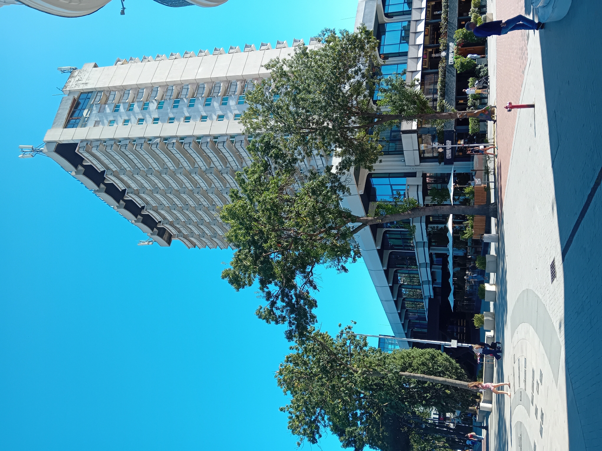 The image captures a sunny day in the city of Varna, Bulgaria, likely near the Varna Congress Center or the Sea Garden. In the foreground, a pedestrian area is paved with light and dark bricks, featuring circular patterns and some embedded square elements. A red fire hydrant stands prominently on the right side of this area.
A group of people are visible walking and standing. On the left, a woman in a light pink dress walks towards the viewer. Further back, another woman pushes a stroller. Several individuals are seen casually walking on the paved area and near the buildings.
The background is dominated by a large, multi-story building with numerous balconies, suggesting a hotel or residential complex. The building has a modern, somewhat Brutalist architectural style, with a light-colored facade. Two trees with dense green foliage partially obscure the lower levels of this building and another structure with large, blue-tinted windows, possibly housing shops or restaurants. These businesses have outdoor seating areas with white umbrellas. In the far distance, a clear blue sky stretches overhead. The overall impression is that of a vibrant urban environment on a pleasant day.