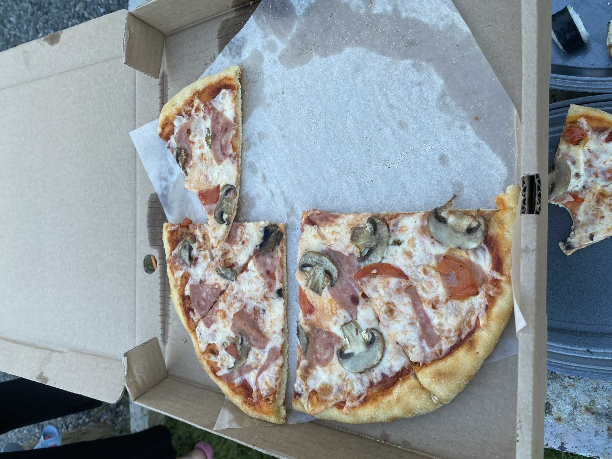 An overhead shot captures a partially eaten pizza in an open cardboard box. The pizza is cut into slices, with some missing. The toppings appear to include cheese, ham, tomatoes, and mushrooms. The pizza is resting on a sheet of parchment paper inside the box. The background shows the edge of the box and what appears to be a paved or gravel surface. The image is taken outdoors, with glimpses of grass and possibly people's legs in the background. The scene does not provide any specific information that would indicate a location in Ufa, Russia.