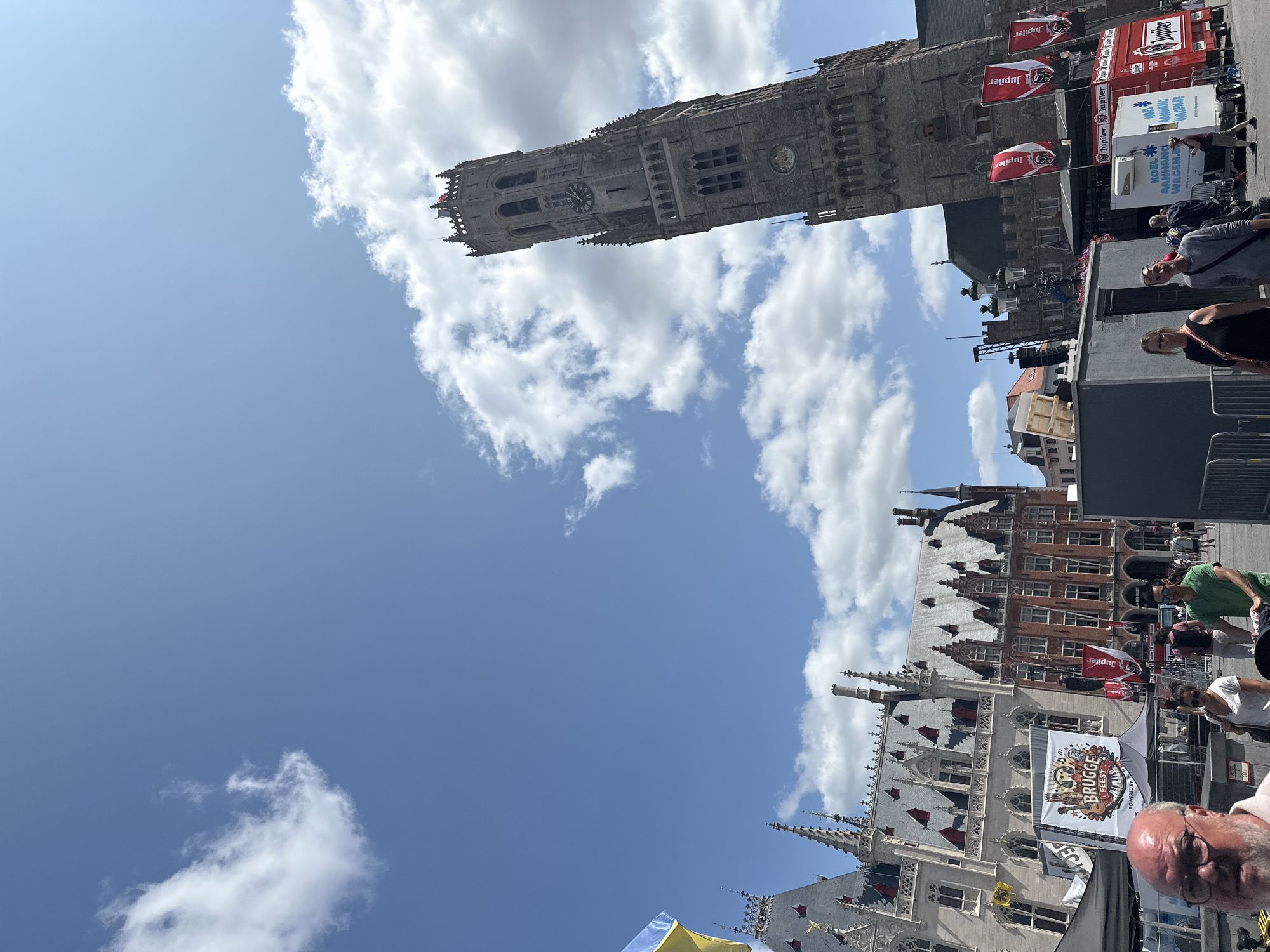 This image captures a view of the Markt square in Bruges, Belgium, featuring the iconic Belfry of Bruges and the historic Provincial Court. The sky is blue with scattered white clouds.
In the foreground, a man with a bald head and glasses is visible on the left, looking towards the camera. Behind him, a crowd of people are walking and standing. Tents and event structures are set up, with banners displaying "BRUGGE FEEST". Flags, including one with the Ukrainian colors, are also present.
The Provincial Court, a Gothic building with ornate spires and red-tiled roofs, stands on the left side of the square. To its right, the imposing Belfry of Bruges, a medieval bell tower, dominates the skyline. The tower has a clock face and is decorated with stone carvings. Red flags with a rose logo are displayed on poles in front of a large red truck with "Jupiler" branding.
The scene appears to be during a daytime event, possibly a festival or market, given the temporary structures and the presence of vendors and attendees. The architecture and the specific landmarks confirm the location as Bruges.