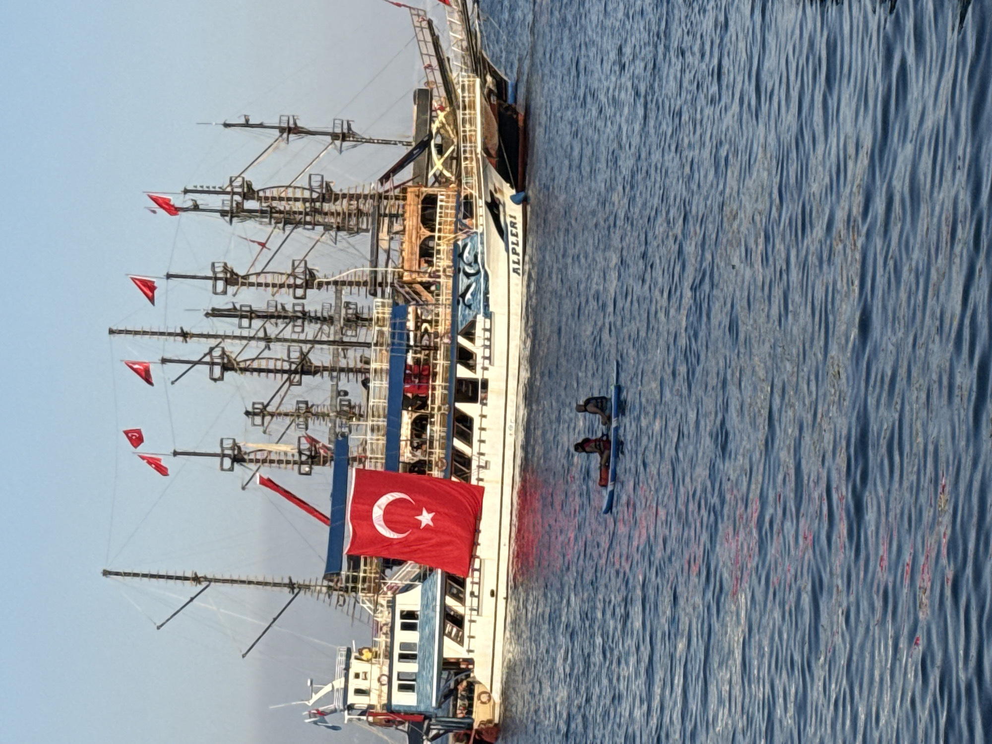 A large white boat with multiple masts and several Turkish flags is docked in the water. The boat has a large Turkish flag draped over its side. In the foreground, two people are paddling on a stand-up paddleboard in the blue water. The water appears calm with slight ripples. The sky is clear and light blue. The boat appears to be a tourist vessel, possibly in Alanya, Turkey, given the Turkish flags and the name "ALP LERI" visible on the side of the boat.