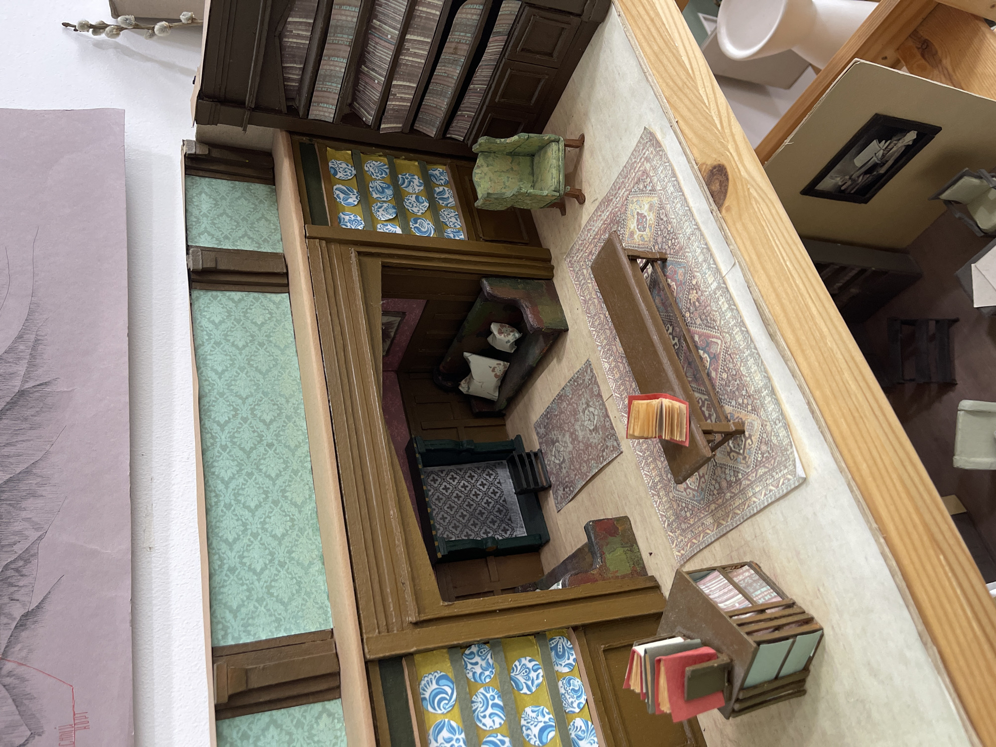 The image shows a detailed dollhouse interior, possibly a representation of a historical or fictional space. The dollhouse is constructed from wood and features multiple rooms, each with meticulously crafted furniture, wallpaper, rugs, and other decorative elements. There is a library with bookshelves filled with tiny books, a living room with a sofa and fireplace, and other rooms with tables, chairs, and decorative objects. The miniature details and craftsmanship suggest this could be a collector's item or a display piece in a museum or private collection. There is no information in the image to confirm its location in Stavropol, Russia, or any specific location.