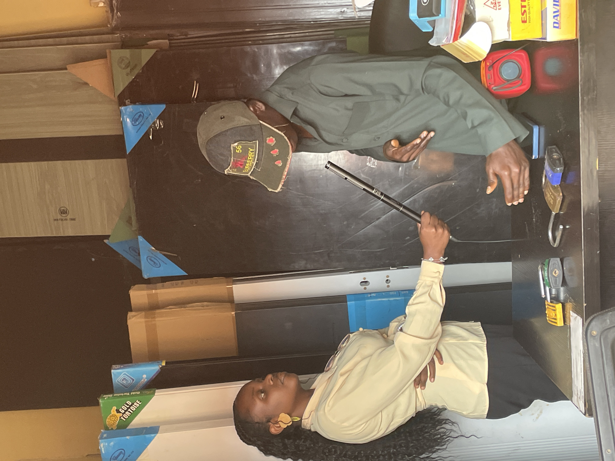 In Kubwa, Nigeria, a woman with dark, curly hair, wearing a light yellow button-down shirt and dark pants, stands on the left side of the frame. She holds a black cylindrical object, possibly a microphone or recording device, extended towards a man on the right. The man, wearing a dark green collared shirt and a dark cap with a patch that reads "BURBERRY 56," is looking down and gesturing with his hands. They are positioned behind a dark countertop on which several items are visible, including two padlocks, a yellow object resembling a lighter, a blue smartphone, a red electronic device, and stacked boxes of various brands. The background features tall, dark objects that appear to be doors or cabinets, along with some cardboard boxes and blue packaging with triangular elements. The environment suggests an indoor setting, possibly a shop or storage area.
