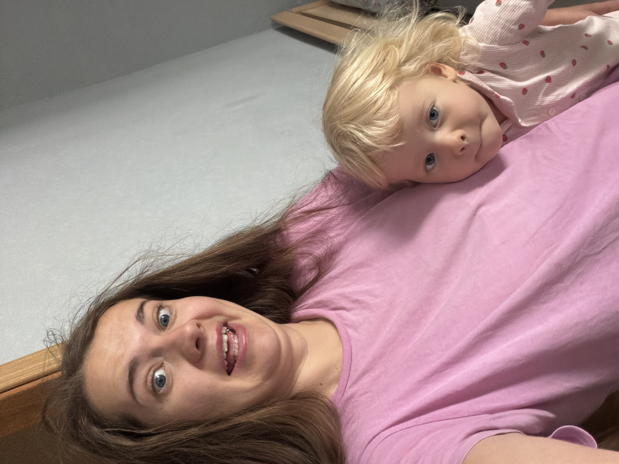 An upward-angled close-up selfie captures an adult female and a young child lying down together, likely in a bedroom in Sochi, Russia. The adult, positioned in the foreground and to the left, has long brown hair and wide blue eyes. She wears a pink t-shirt and displays a playful or surprised expression, with her mouth open and tongue slightly visible.
Resting her head on the adult's chest or shoulder, positioned to the right and slightly above the adult, is a young child with blonde, slightly curly hair and blue eyes. The child is wearing a light pink and white striped top adorned with small red strawberry patterns. Her gaze is directed towards the camera with a calm, slightly curious expression.
The background reveals a light grey sheet or bed surface, with a wooden headboard or bed frame visible on the left side. The image conveys a casual, intimate, and playful moment between the two individuals. There is no visible text.