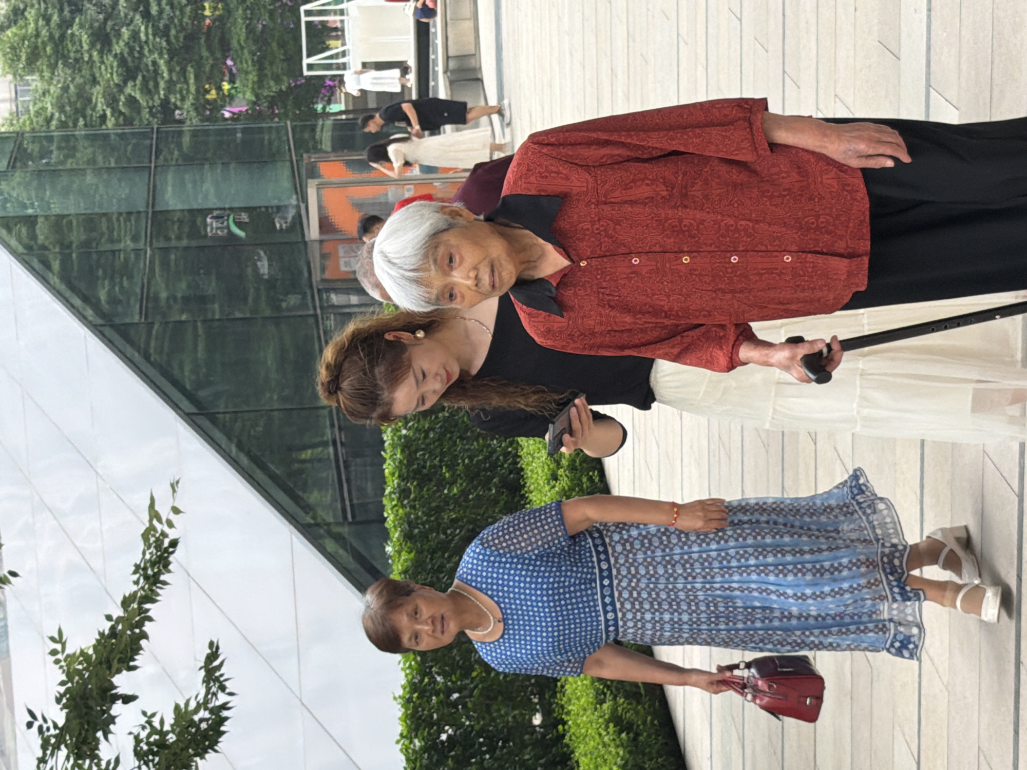 Three women are walking on a tiled outdoor walkway in front of a modern building with a glass facade and a sloping roof. The woman on the left is wearing a blue and white patterned dress, a pearl necklace, and white shoes, and is carrying a red handbag. The woman in the middle, with her hair in a bun, is wearing a black top and a long white skirt, and is looking at her mobile phone. The woman on the right, who appears to be elderly, has white hair and is wearing a red patterned shirt and black pants. She is holding a black cane in her right hand. In the background, other people are visible walking and standing, and trees and foliage line the walkway. The scene appears to be in a public space within the city of Yuzhong District, China.
