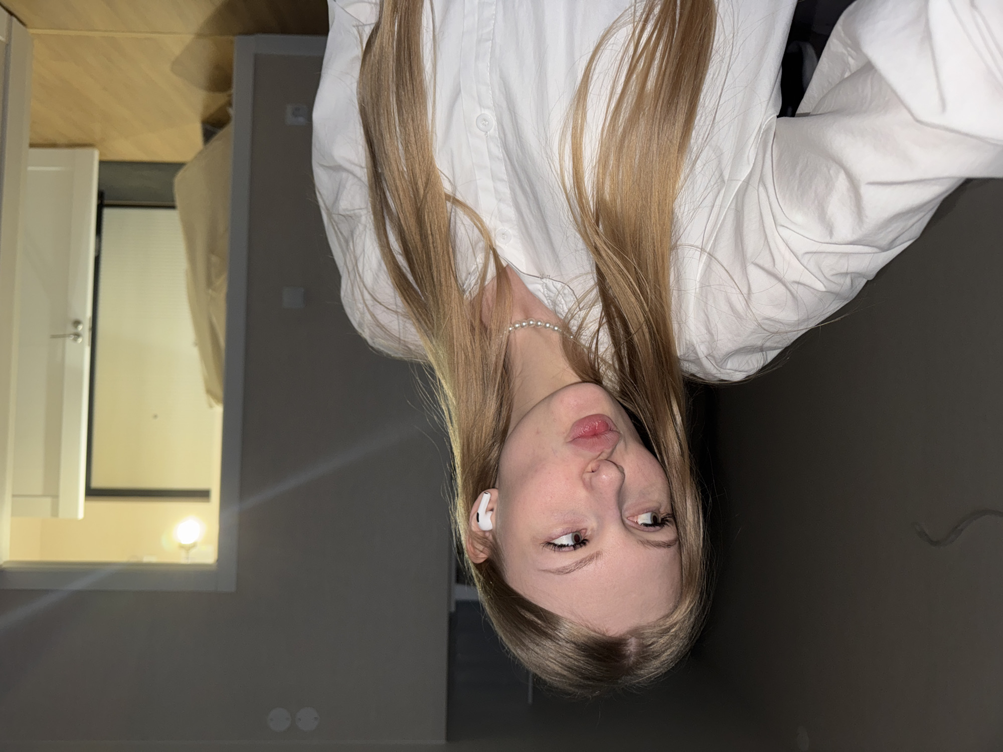 A full-shot portrait features a young woman with long, straight blonde hair, fair skin, and blue eyes, captured indoors. The woman is wearing a white button-up shirt and a white pearl bracelet on her left wrist. A white earbud is visible in her left ear. Her head is tilted slightly downwards and to the side, with her eyes looking upwards and to the left, and her lips gently pursed in a slight pout.
The setting is an indoor room with light-colored, possibly beige or light grey, walls. In the background, to the left, a white door with a handle is visible. Further back, an opening or window reveals a bright light source, possibly a lamp from an adjacent room or a light outside, casting a glow. The floor appears to be made of light-colored wood. Two circular fixtures, possibly electrical outlets or light switches, are visible on the lower part of the wall. The ambient lighting suggests it might be evening or nighttime. There is no visible text in the image. The overall impression is a casual, posed portrait.