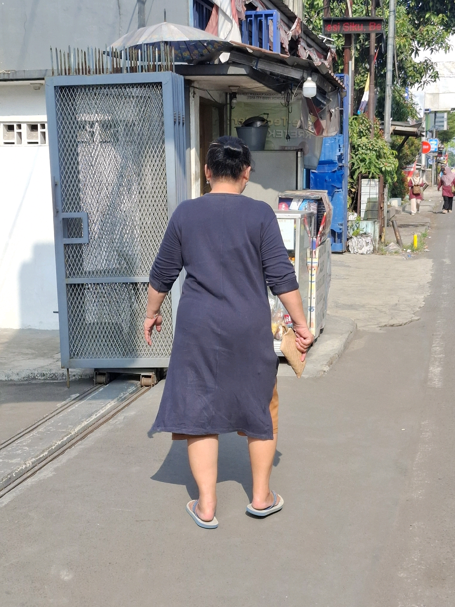 The image depicts a person walking away from the camera on a paved street in Sukaraja, Indonesia, on a sunny day. The person is wearing a dark blue, long-sleeved dress that reaches below the knees, and light blue flip-flops. They are holding a cone-shaped object, possibly an ice cream cone, in their right hand. To the left of the person is a grey metal gate with diamond-shaped mesh, and a white wall with some vents. To the right, there is a small storefront with a blue awning and a display case. Behind the storefront, several people are walking on a sidewalk, and trees and utility poles are visible. A digital sign with text is mounted on a pole above the storefront. The street appears to be a commercial or residential area, with buildings and businesses lining the road. The overall scene suggests a casual, everyday moment in a bustling Indonesian city.