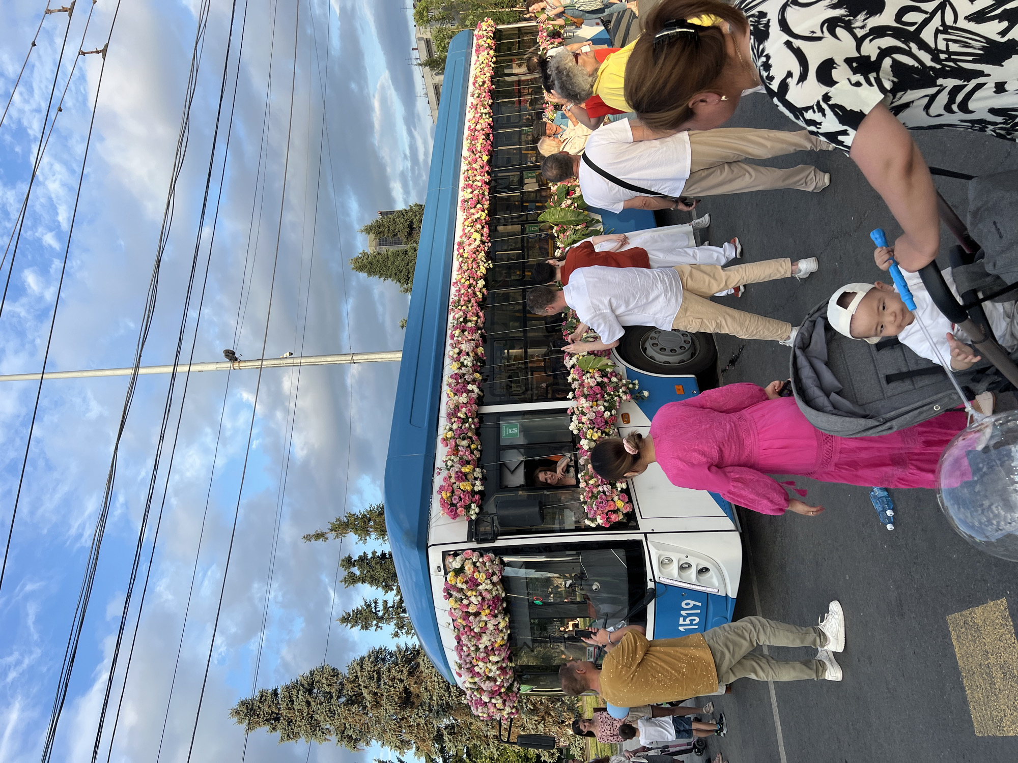 This media file captures a decorated bus, likely for an event or celebration, in Novosibirsk, Russia. The bus is adorned with numerous colorful flowers, primarily pink and yellow, along its windows and front. Several people are gathered around the bus, some appearing to be passengers and others onlookers.
In the foreground, a man in a yellow shirt is holding a phone, possibly taking a photo or video. Next to him, a woman in a patterned black and white top is looking down. A baby in a stroller, wearing a white cap and onesie, is also visible. Further down the bus, a woman in a vibrant pink dress stands by the side. People in white shirts and khaki pants are also seen near the bus.
The scene appears to be outdoors during daylight, with a partly cloudy sky. Trees line the background, and power lines are visible overhead, suggesting an urban or suburban setting. The overall atmosphere seems festive, with people engaged in observing or participating in whatever event is occurring. The number "1519" is visible on the side of the bus.