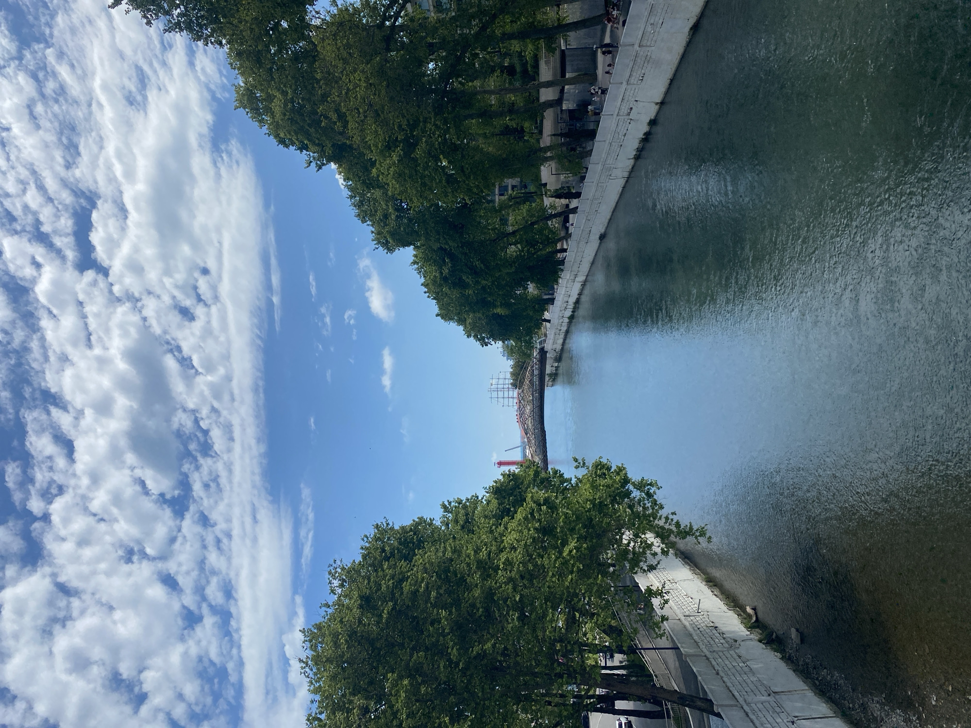 The image depicts a view of the Sochi River in Sochi, Russia, on a partly cloudy day. The river flows through a channel with concrete retaining walls on both sides. Lush green trees line the banks, with some buildings visible in the background. In the distance, a bridge spans the river, and a red lighthouse is visible near the bridge. Several people are seen walking and sitting along the riverbanks. The water is calm with reflections of the sky and surrounding trees.