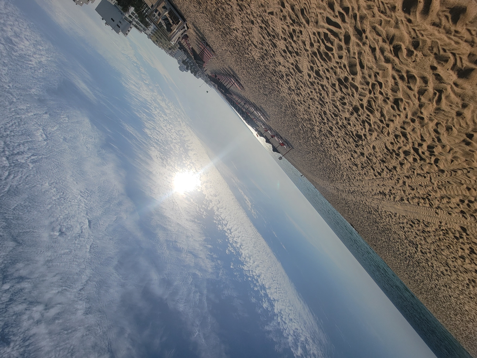 The image depicts a beach scene in Quarteira, Portugal, oriented sideways. The foreground is dominated by sand dunes, with tire tracks visible. In the middle ground, rows of red sun loungers and umbrellas are arranged on the beach. Beyond the loungers, buildings line the coast, suggesting a developed resort area. The sea is visible in the distance, and the sky is filled with scattered white clouds. The sun is shining brightly, creating a glare on the water and casting shadows on the sand, indicating it is daytime. The overall impression is one of a bright, sunny day at a popular beach destination.