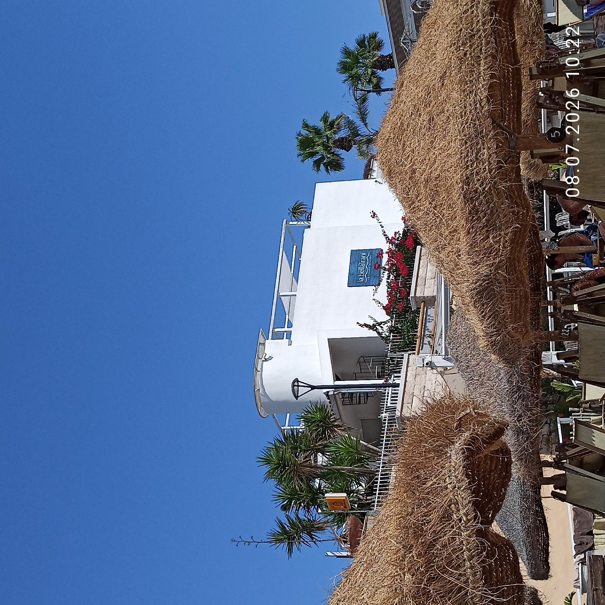 The image shows a bright, sunny day at a beachside establishment, likely in Trani, Italy, as indicated by the presence of palm trees and the clear blue sky. In the foreground, several thatched umbrellas provide shade over beach chairs, suggesting a relaxed vacation setting. Behind the umbrellas, a white building with a modern, minimalist design is visible, featuring a curved section, a spiral staircase, and balconies. A prominent blue sign on the building reads "la bella Trani". Lush green plants, including palm trees and flowering bushes with red blossoms, surround the building, adding to the Mediterranean ambiance. The image also includes a timestamp in the bottom right corner: "08.07.2026 10:22".