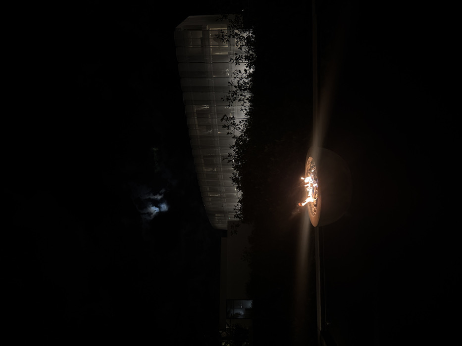 At night, a glowing fire pit is on the right. A tall building with linear, grid-like windows is on the left, partially obscured by dark foliage. A hazy, illuminated moon is visible behind dark clouds in the background. The scene is dark, with the fire pit and building casting the only significant light.