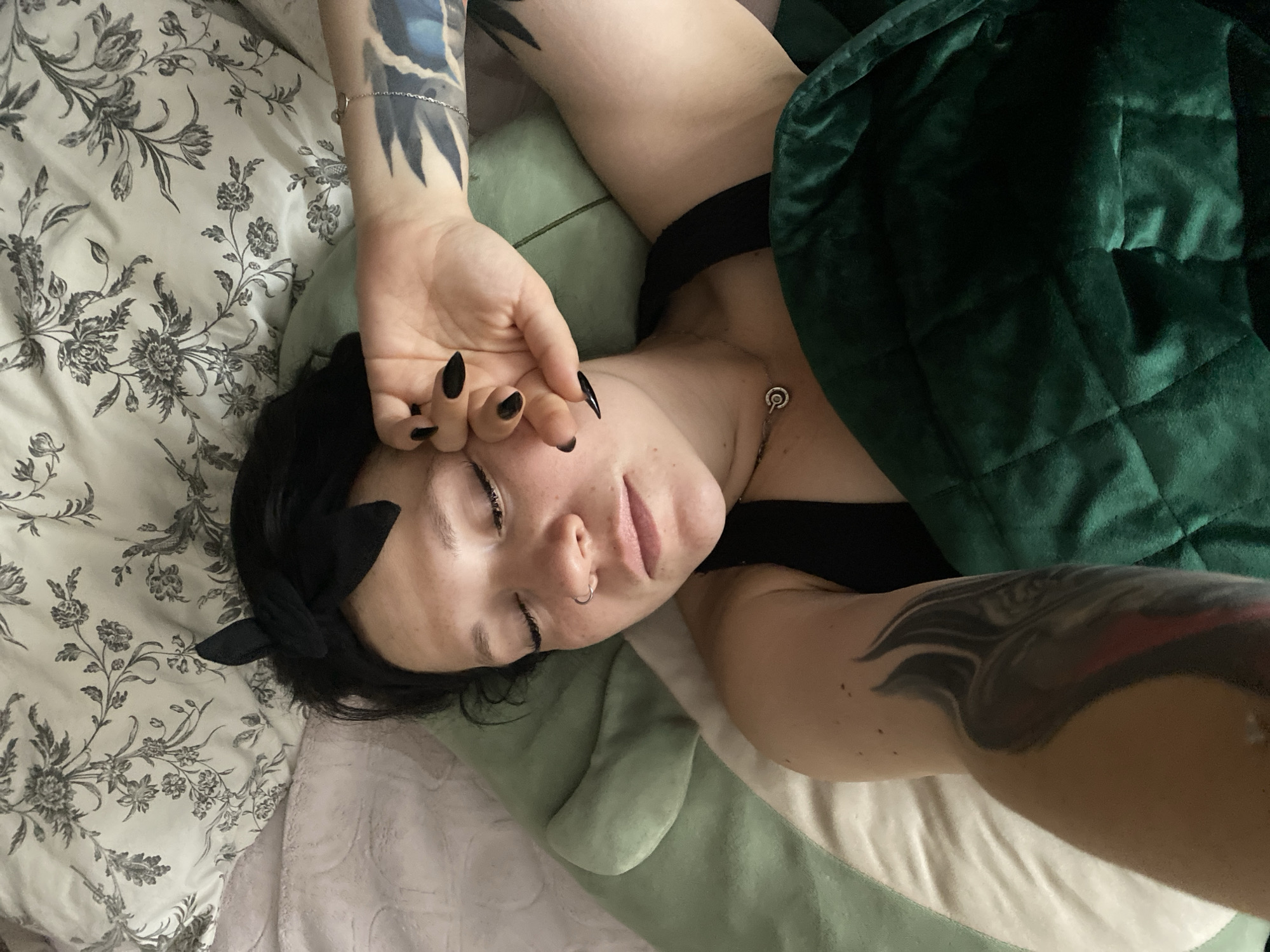 A young woman with dark hair and a black headband is lying in bed, eyes closed, with her right hand resting on her face. She is wearing a black tank top and a silver necklace. She has tattoos on her left arm and right arm. The bed is covered with a green blanket and floral patterned sheets. The scene is indoors, likely a bedroom, with no visible indication of the city of Chaikovsky, Russia.