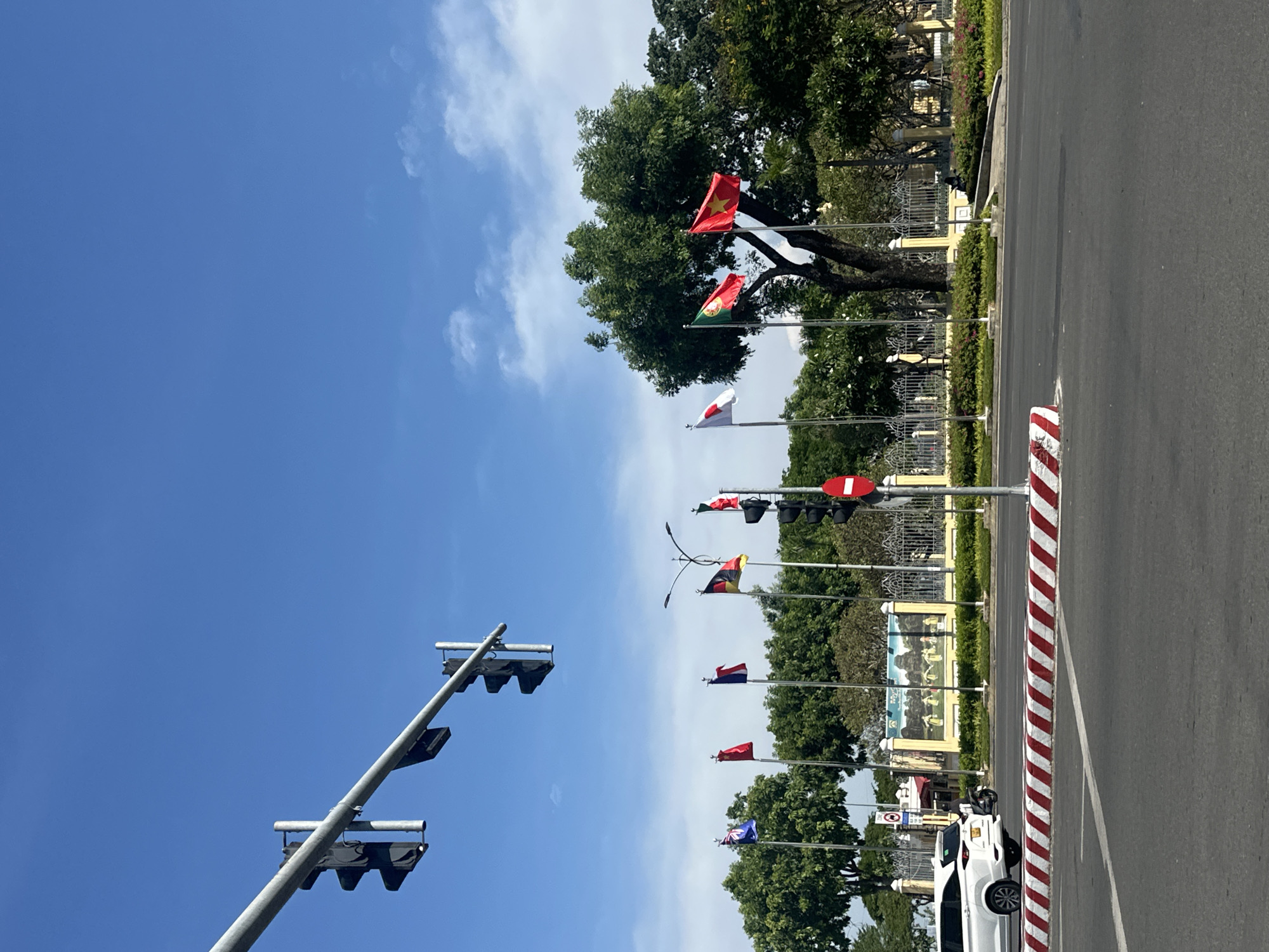 This media file captures a bright, clear day in Đà Nẵng, Vietnam, likely mid-morning or early afternoon given the strong sunlight and blue sky with scattered white clouds. The scene is a city street with flags displayed prominently along the roadside.
The primary subjects are a series of national flags mounted on poles, interspersed with traffic lights. Visible flags appear to include Vietnam's red flag with a yellow star, Japan's flag with a red circle, and several others in various colors and designs, suggesting an international or governmental setting. The flags are positioned along a well-maintained street, with a paved road and a striped barrier delineating the traffic lane from a sidewalk or median.
In the background, lush green trees form a canopy, partially obscuring buildings that appear to be ornate, possibly government structures or historical landmarks, with yellow accents. A large mural or artwork is visible on a wall behind the flags.
The activity taking place is simply the display of flags, likely for a public event, diplomatic occasion, or as a general display of national pride. There are no people clearly visible in the immediate foreground or engaged in any specific activity, though a white car is parked on the side of the road. The overall impression is one of order and civic display on a pleasant day. The traffic lights are functional, with some showing red signals, indicating normal traffic flow. The image is oriented vertically, with the sky occupying the upper portion of the frame and the street scene extending downwards.