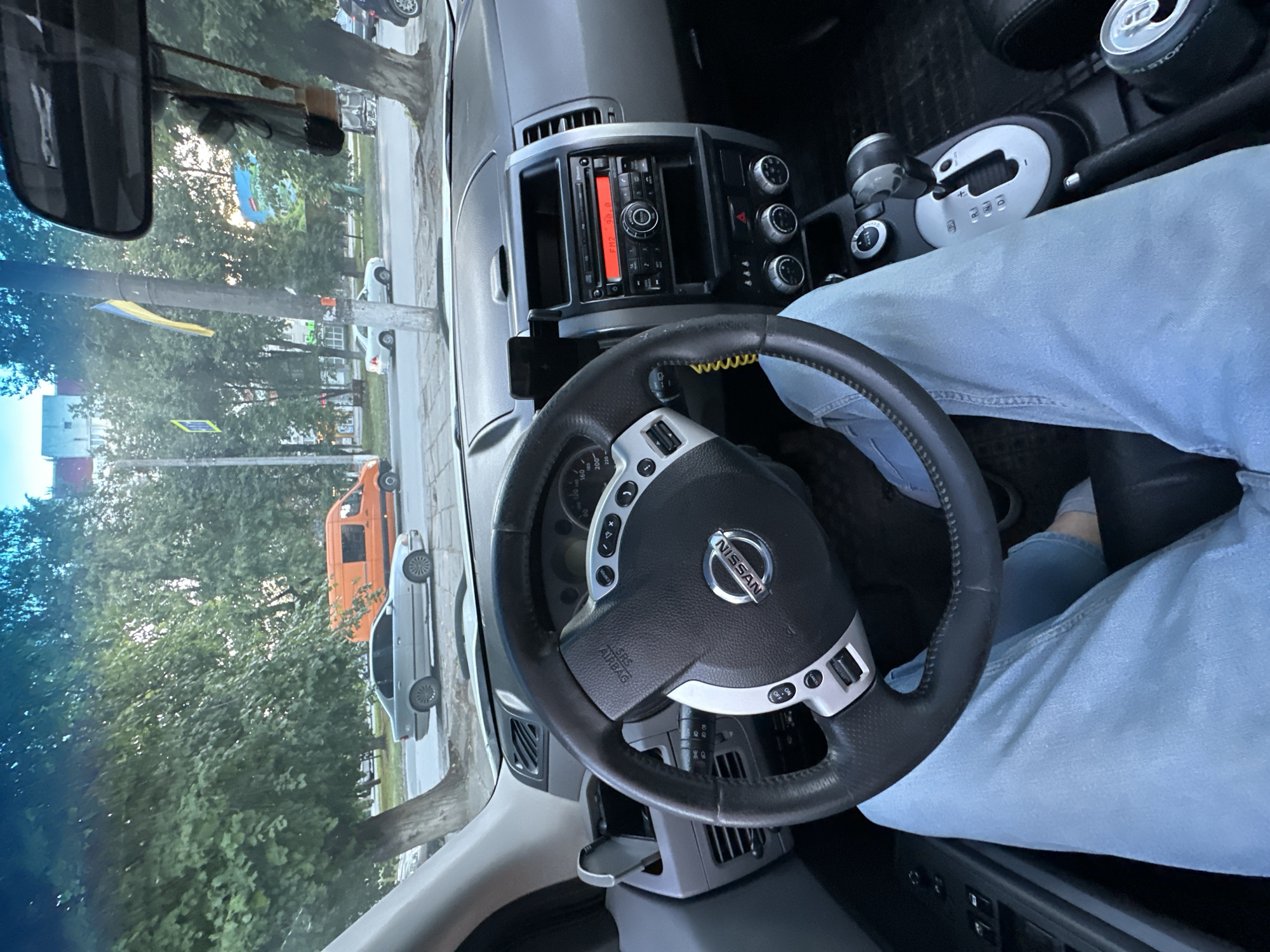 The image is a first-person view from inside a Nissan SUV, looking forward through the windshield. The driver, wearing light blue jeans, has their hands on the steering wheel and their legs visible in the foreground. The steering wheel has a Nissan logo in the center and controls on the sides. The dashboard features an infotainment system with a red display showing "FM2 88.8", climate control knobs, and a gear shifter. Outside the car, through the windshield, a street scene is visible. Cars, including a silver sedan and an orange van, are parked or driving on a paved road. Trees line the street, and buildings are visible in the background. A Ukrainian flag hangs from a utility pole. The scene appears to be in an urban or suburban environment, possibly during daylight hours, with a slightly overcast sky. The composition suggests the vehicle is either parked or moving slowly. The general setting is consistent with a street in Kamianets-Podilskyi, Ukraine, given the presence of the Ukrainian flag and typical urban architecture.