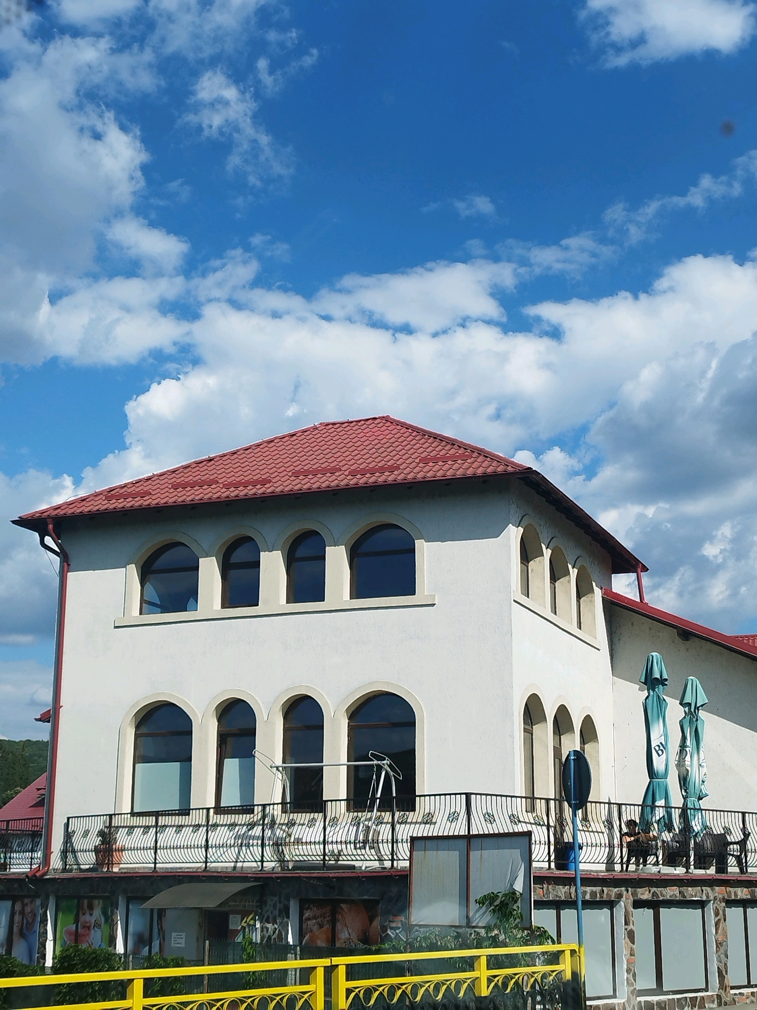 The image shows a white building with a red tiled roof under a bright blue sky with scattered white clouds. The building has arched windows on the upper floors and a balcony with a black wrought-iron railing. The lower part of the building is clad in stone. On the balcony, there are two closed green umbrellas, some chairs, and a person seated. In the foreground, a bright yellow railing runs horizontally across the bottom of the image, obscuring some of the ground level of the building which appears to be a commercial establishment with large windows displaying images. The building is located in Curtea de Argeș, Romania.