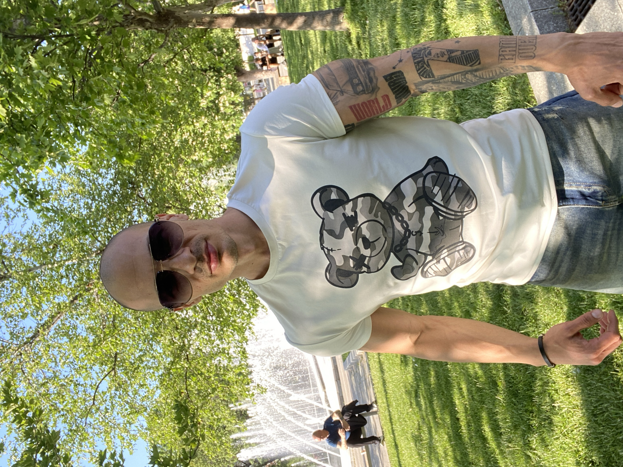 A bald man with sunglasses is posing for a photo in a park in Pleven, Bulgaria. He is wearing a white t-shirt with a gray and black bear design and blue jeans. He has tattoos on his left arm. Behind him is a fountain with water spraying, and two people are walking behind the fountain. The park has trees and green grass. Other people are visible in the background.