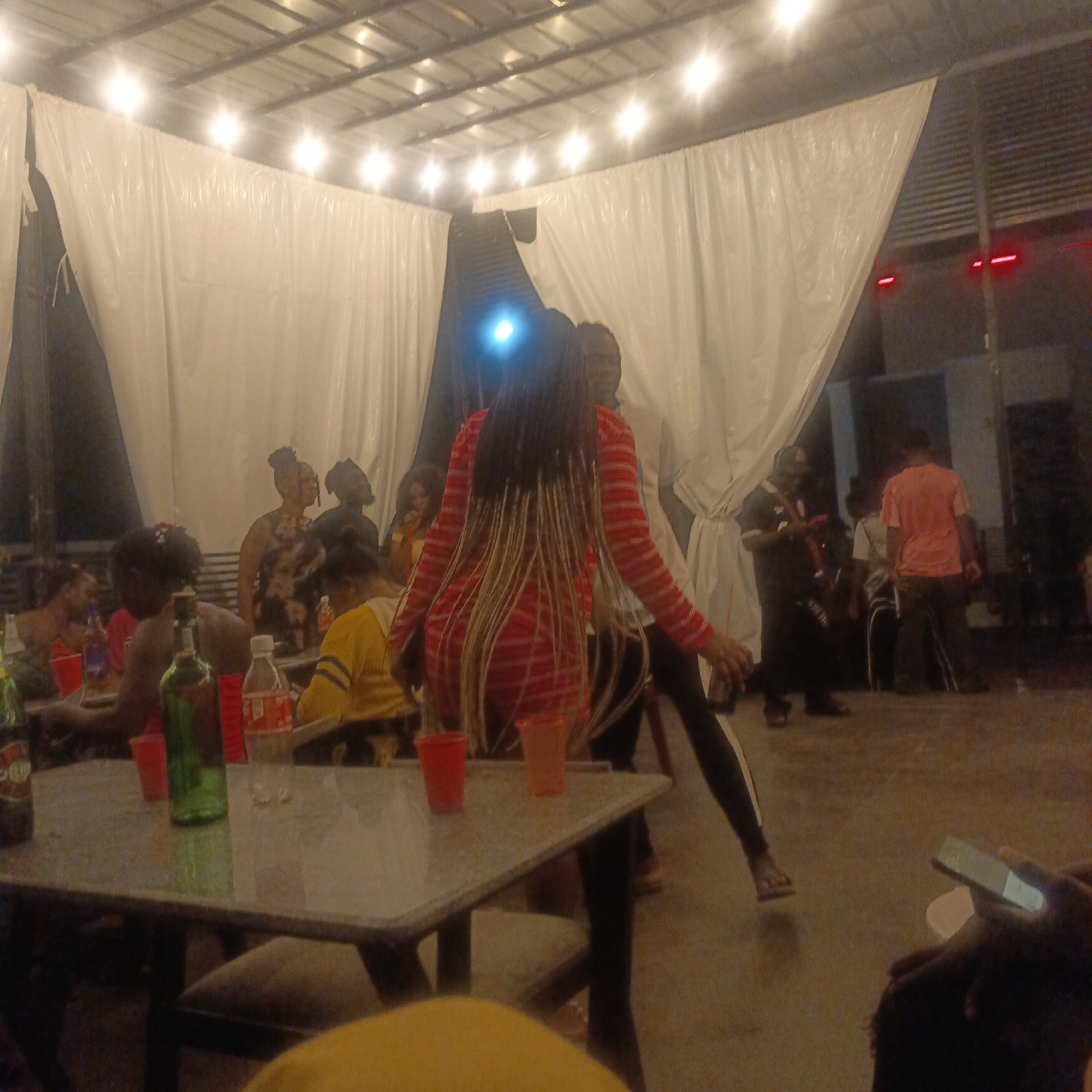 The image depicts a gathering of people at night, likely at an event or party. The location is possibly in the city of Ube, Nigeria.
**People:** There are several people visible. Some are seated at tables, possibly eating or drinking. Others are standing and appear to be dancing or interacting. One person is in the foreground with their back to the camera, wearing a red and white striped dress. Their hair is braided with blonde highlights.
**Objects:** There are tables and chairs visible. There are several green beer bottles on the table in the foreground, as well as red cups. A mobile phone is visible in the lower right of the image, being held by a person.
**Actions:** People are seated and standing, some are dancing. People appear to be eating, drinking and interacting.
**Interactions:** People are socializing.
**Environment:** The scene takes place indoors under a metallic roof with multiple lights. There are white curtains or drapes around the space, giving the impression of a temporary venue.