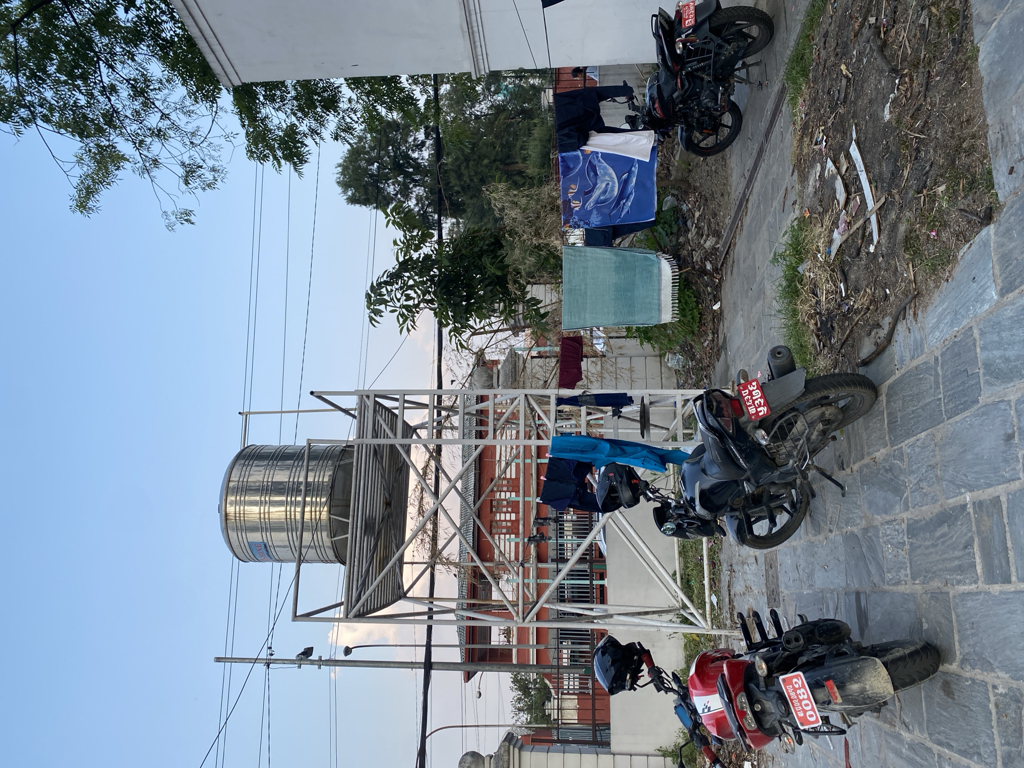 In Kathmandu Metropolitan City, Nepal, the scene depicts a water tank elevated on a metal structure. Below, two motorcycles are parked on a stone-paved area. Laundry is hung to dry on a clothesline. The environment appears to be an urban setting with a building and trees visible in the background, under a clear sky.
