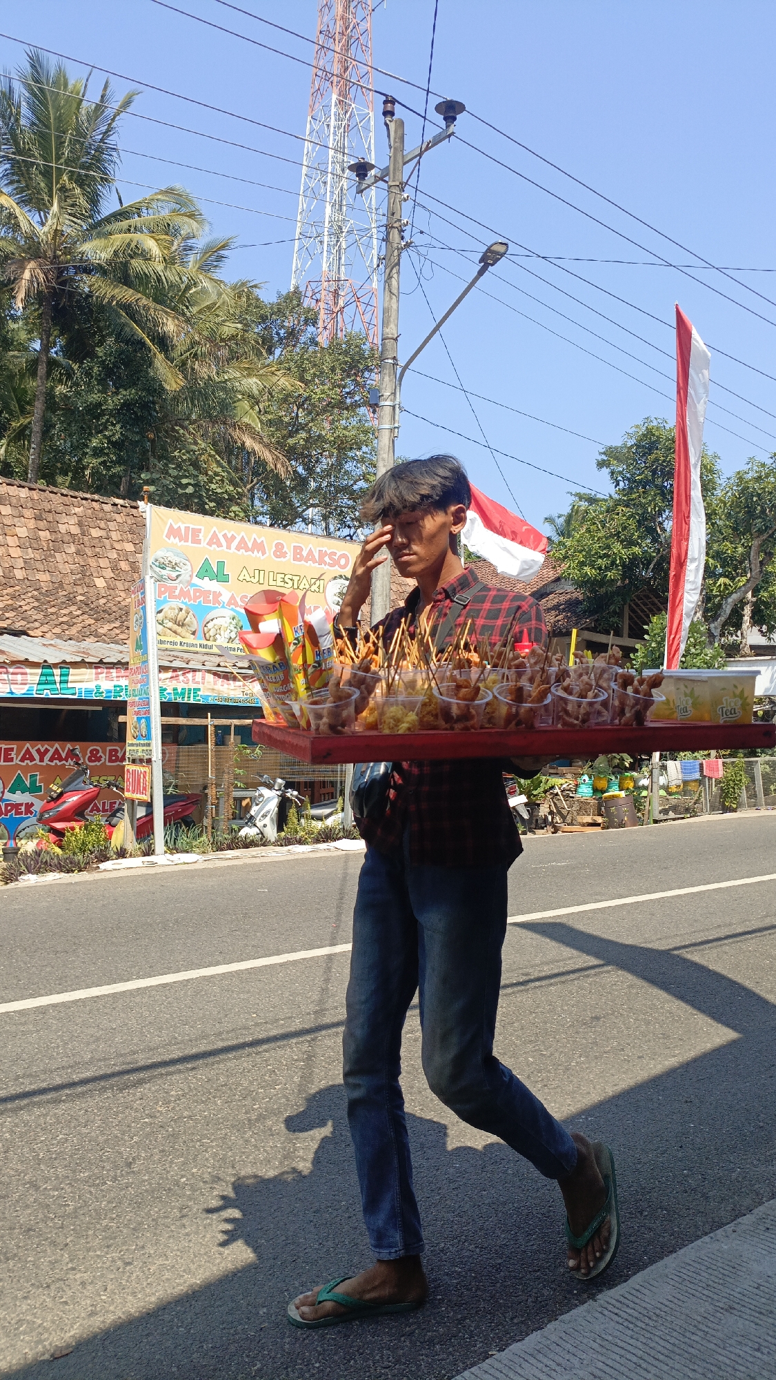A young male vendor walks on the side of a street in Krajan Lor, Indonesia, carrying a red tray laden with food items. The vendor, dressed in a red and black plaid shirt and blue jeans, is holding his hand to his forehead, possibly due to the sun. The tray holds numerous small plastic cups filled with food, each topped with a skewer, and some containers of drinks. In the background, a building with a tiled roof and a banner advertising "MIE AYAM & BAKSO AL AJI LESTARI" is visible. A red and white Indonesian flag hangs on a pole. Further in the background, a tall transmission tower stands against a clear blue sky. The street is paved with asphalt and has a white line marking the lane. Palm trees and other greenery are present on the side of the road. A motorcycle is parked near the building. The overall scene depicts a typical street-side vendor in an Indonesian setting.
