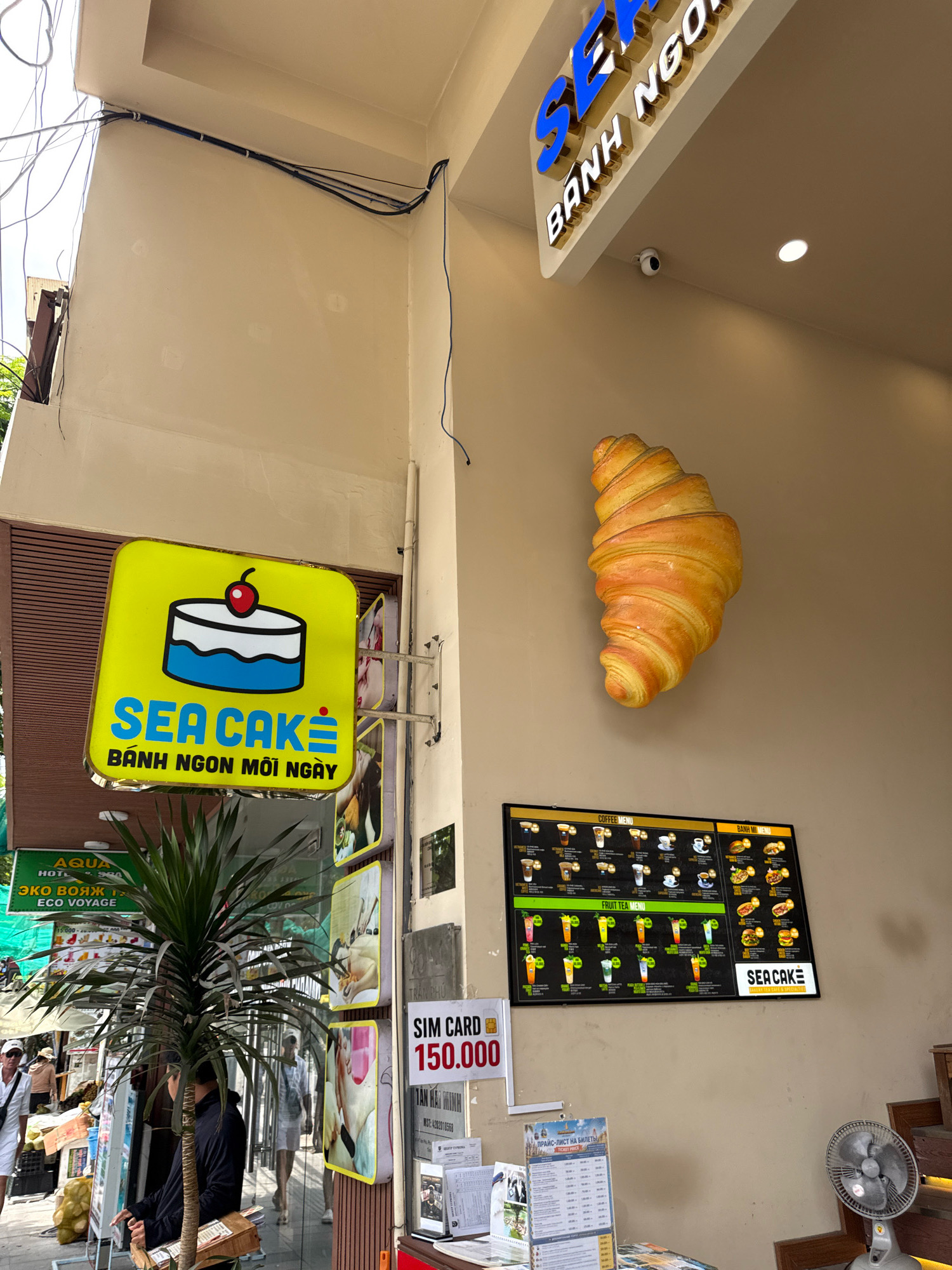 The image captures a street-level view of a cafe or bakery in Nha Trang, Vietnam. A prominent sign for "SEA CAKE" with a cake illustration is mounted on a wall. Above it, another sign reads "SEA BANH NGO". A large, realistic croissant sculpture is affixed to the building. A menu board displaying coffee, fruit tea, and pastry options is visible. To the left, a lush green plant stands in front of a storefront with various signs, including one in Russian that appears to be related to travel ("ECO VOYAGE"). A "SIM CARD 150.000" sign is also present. Several individuals are in the background, some appearing to be customers or staff. The overall environment suggests a bustling commercial area with an outdoor seating or service area.