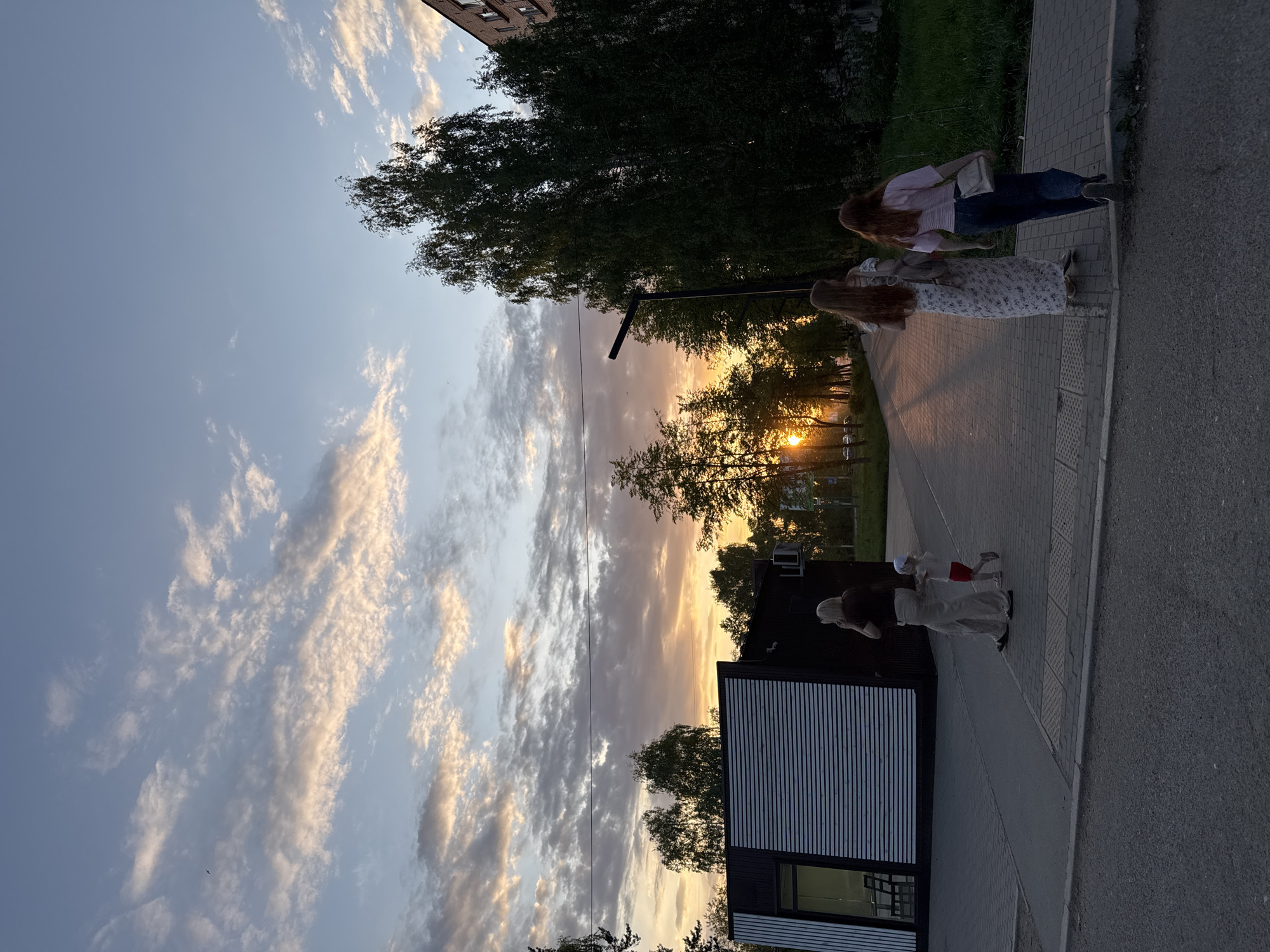 The image depicts a scene in Chaikovsky, Russia, at dusk or sunset. The sky is filled with clouds illuminated by the setting sun, creating a gradient of colors from blue to orange. Two women are walking on a paved sidewalk adjacent to a street. One is wearing a light-colored dress with a floral pattern, while the other is wearing a pink shirt and dark jeans. A building with a striped wall is visible on one side of the sidewalk. In the distance, the sun is setting between trees. The overall impression is one of a peaceful evening stroll.
