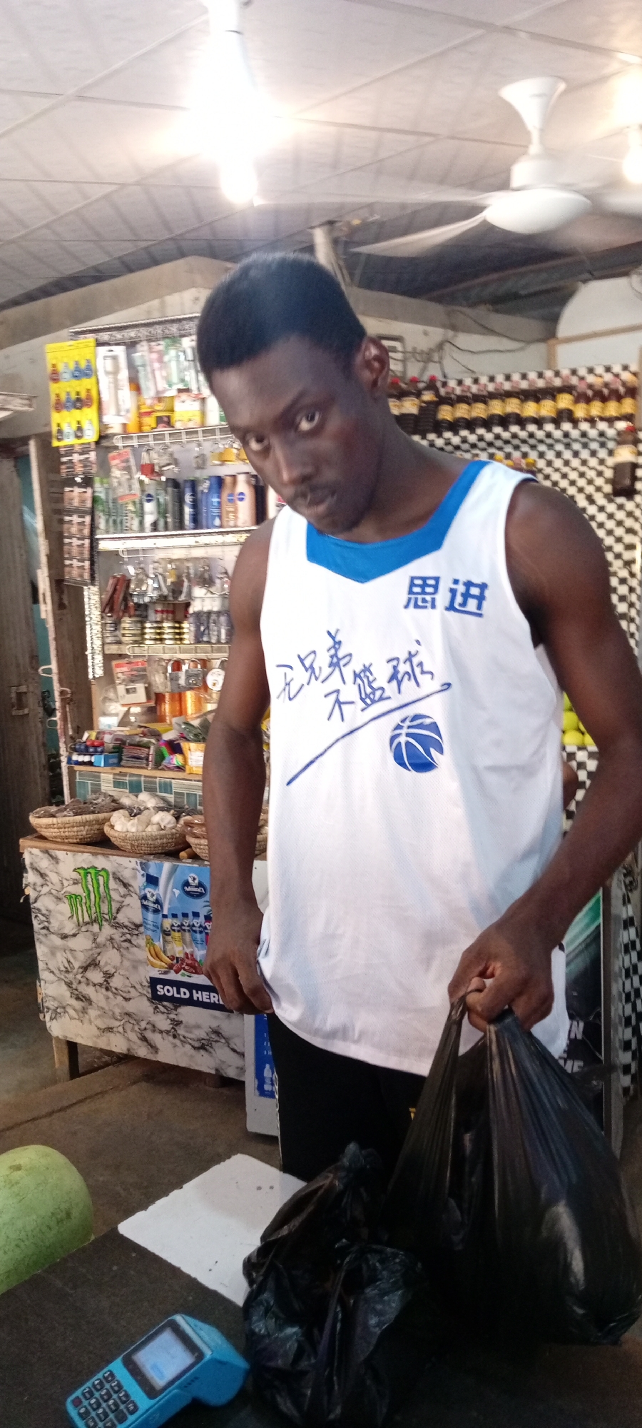 A Black male in a white tank top with blue trim and Chinese characters, stands in front of a small market stall. The tank top has a basketball graphic. He is holding two black plastic bags, one in his right hand and another smaller one at his feet. To his left are shelves stocked with various goods including bottles of liquid, packaged items, and baskets of goods. Behind him, more shelves and a checkered pattern are visible. In the foreground, a blue card reader and a large green fruit are on a counter. The scene appears to be an outdoor market in Aviation Quarters, Nigeria.