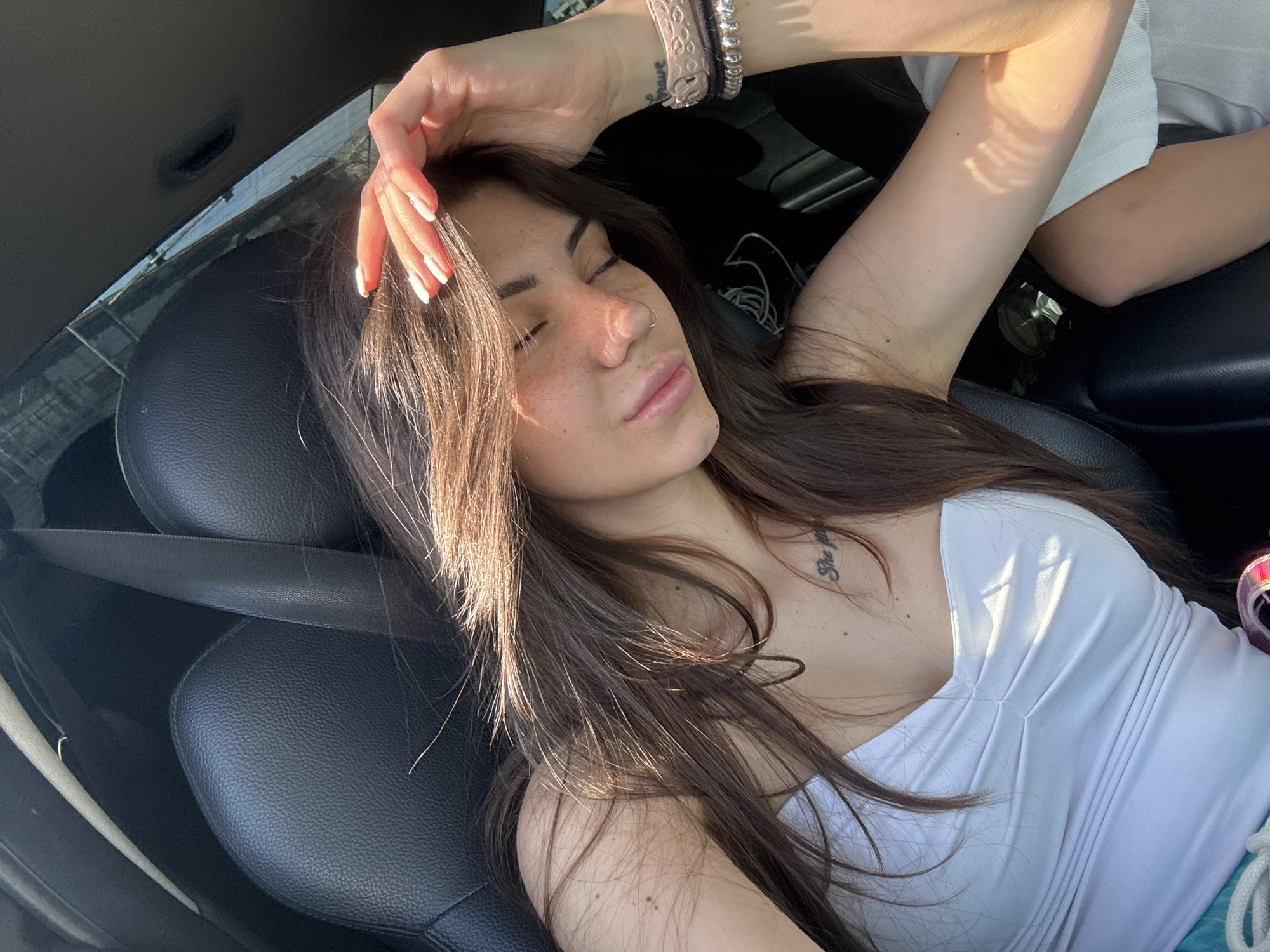 A young woman with long brown hair and closed eyes leans back on a black leather car seat. She is wearing a white tank top and has a tattoo on her chest. Sunlight streams in, illuminating her face and arm. Her right arm is raised, with her hand resting on her forehead. Bracelets are visible on her left wrist. The background shows the interior of a car, with a seatbelt across the frame. The image appears to have been taken during daylight hours. The location is unspecified, but it is likely a vehicle.