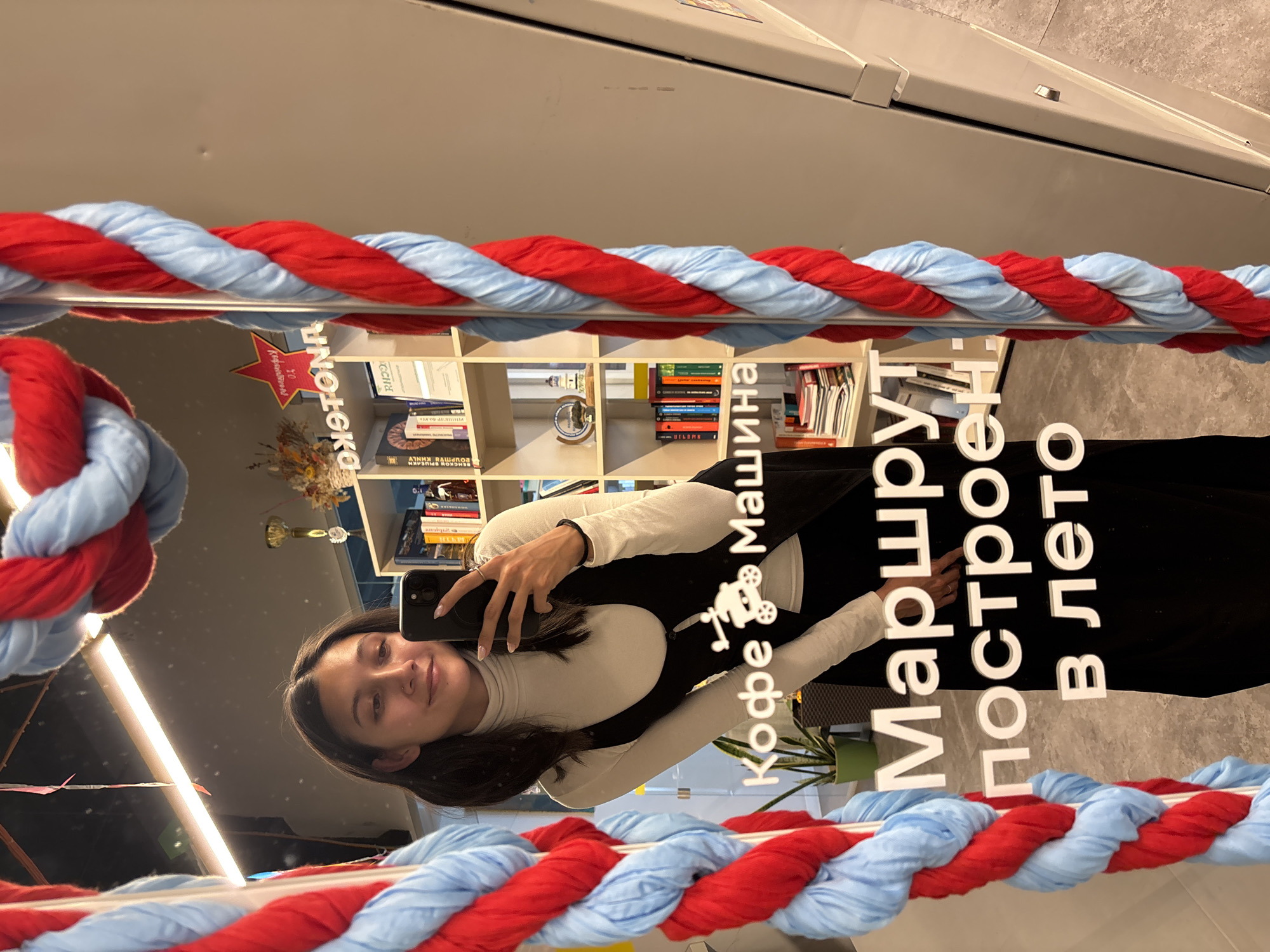 The image, originally rotated, shows a young woman taking a mirror selfie indoors. She is reflected in a large mirror, smiling gently at the camera held in her right hand. She has dark hair and is wearing a light-colored turtleneck top under a black sleeveless garment or dress.
The mirror itself is bordered on the top and bottom by a decorative twisted rope or garland, alternating in red and light blue colors. The background, also reflected in the mirror, consists of several white bookshelves filled with numerous books, indicating a setting like a library, bookstore, or a cafe with a literary theme. Some decorative items, including what appears to be a vase with dried flowers and a potted green plant, are also visible on the shelves. The floor visible in the reflection is a light grey.
Several lines of white Russian text are prominently displayed on the mirror's surface. Reading from top to bottom, the text includes:
1. "КИНОТЕКА" (KINOTEKA), meaning "Cinematheque" or "Film Library," accompanied by a red star logo that reads "Вход свободен" (Vkhod svoboden), meaning "Free entry."
2. "Кофе Машина" (Kofe Mashina), meaning "Coffee Machine."
3. "Маршрут построен в лето" (Marshrut postroyen v leto), which translates to "The route is laid out for summer" or "The route is built into summer."
This text suggests the location is a cultural or commercial space in Vladivostok, possibly combining elements of a film library/cinema with a coffee shop, perhaps with a seasonal or travel-related promotional theme for summer. The overall mood is calm and inviting.