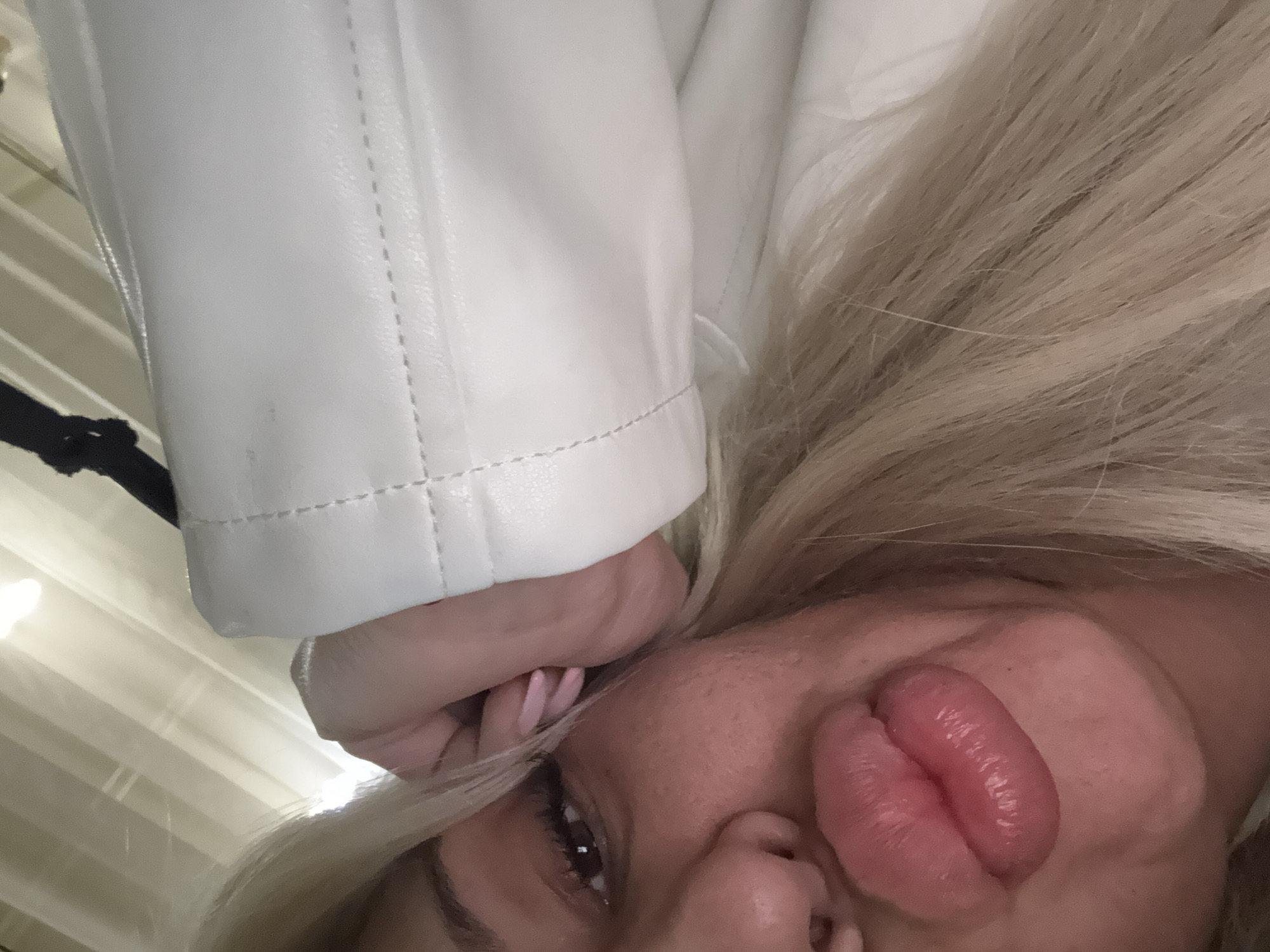 This rotated close-up image, set in Plovdiv, Bulgaria, features a person, likely a woman, with long blonde hair. Her face occupies the lower right portion of the frame, showcasing one eye, part of her nose, and her prominent, puckered lips in an exaggerated "fish face" or kissing pose. Her lips appear full and glossy. A hand, possibly her right, with light pink nail polish, is visible near her face.
She is wearing a white garment, likely a jacket or coat, made of a material resembling leather, with visible stitching details on the sleeve and body. Her blonde hair flows around her face and shoulder. The background consists of light-colored vertical blinds or panels, suggesting an indoor setting with even, soft lighting. A dark strap or cord is visible in the upper left corner. The image conveys a playful and casual tone, likely a selfie or a close-up portrait. There is no visible text.