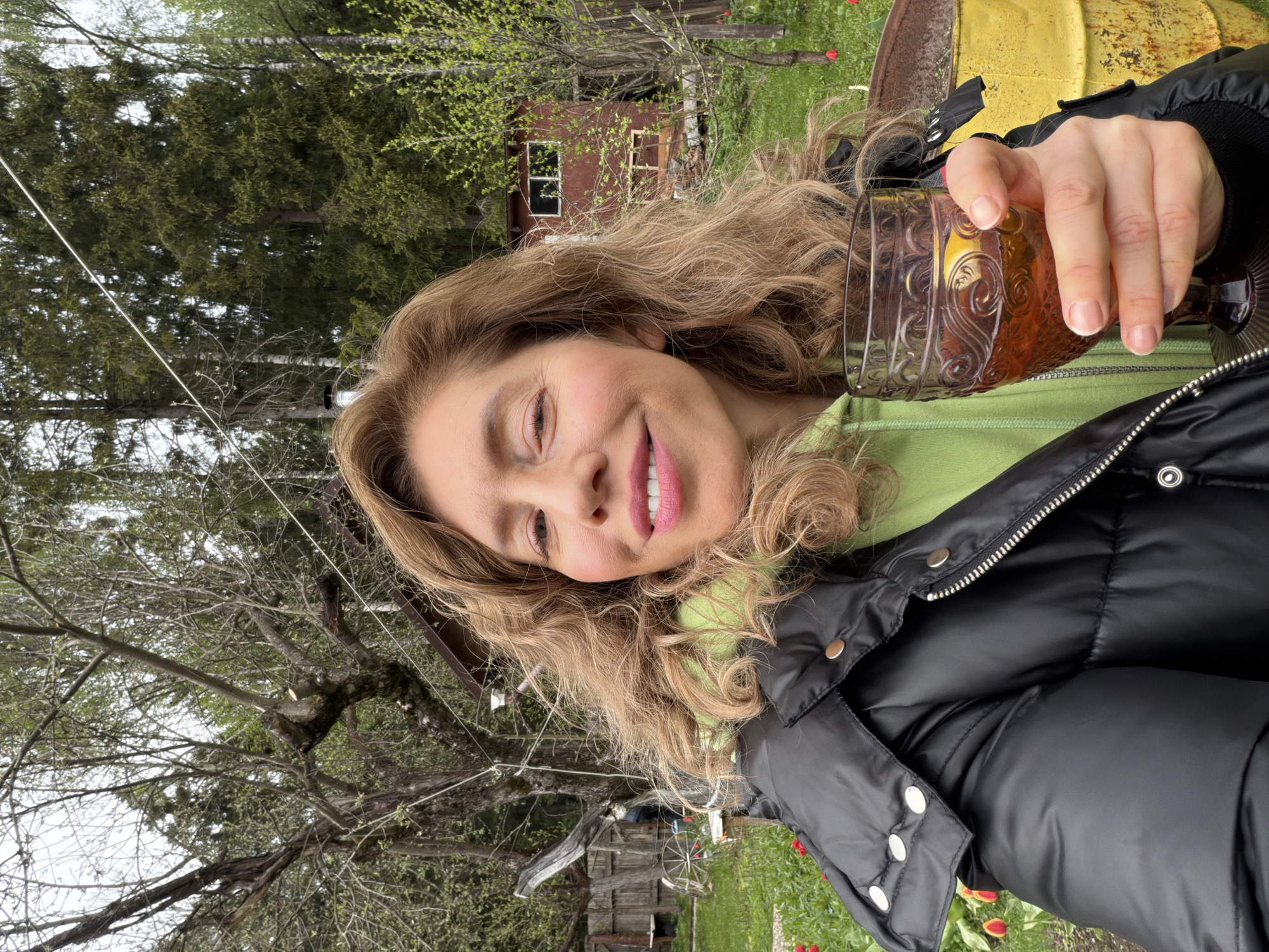 A woman with blonde curly hair and fair skin, wearing a light green hooded sweatshirt and a black puffy jacket with silver buttons, is holding an ornate glass filled with a brown liquid. She is smiling, facing towards the camera at an angle. Behind her, there are trees, a wooden shed, and a building with a window. There are also red flowers, possibly tulips, visible in the grass. A partially visible rusted metal container, painted yellow, is also in the background. The location appears to be a rural outdoor setting.