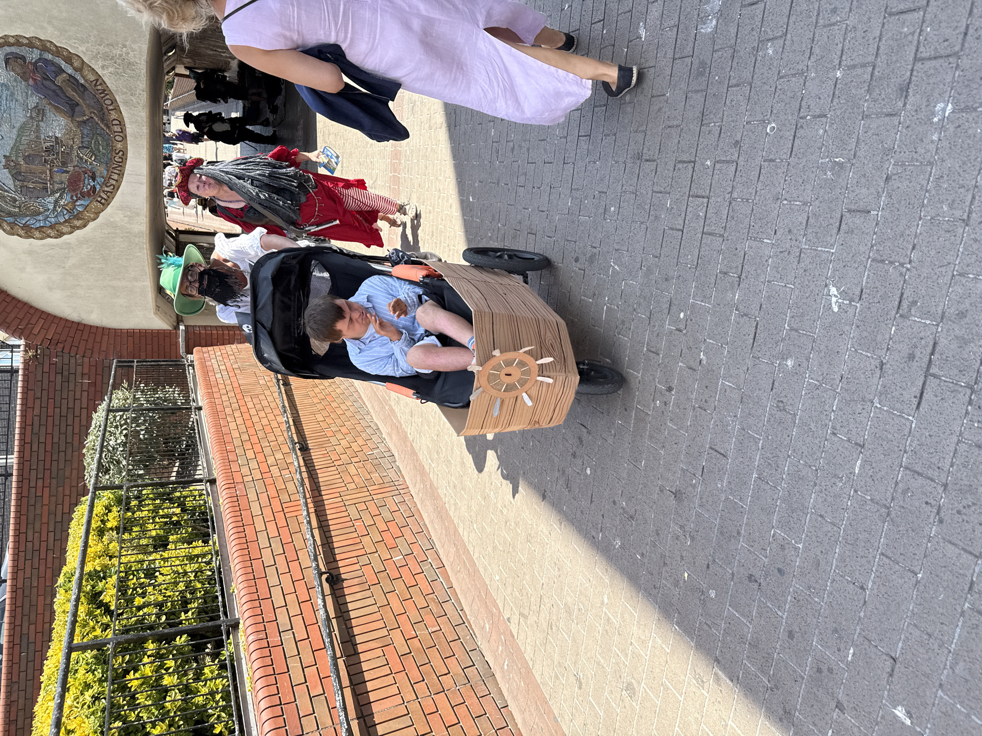 A person with Down syndrome sits in a custom-made stroller shaped like a wooden ship's barrel, wearing a light blue striped shirt and white shorts, facing forward and looking down, possibly eating. The stroller has a wooden ship's wheel attached to the front. Two adults in pirate costumes, one with a green hat and beard, and another with a red bandana and grey dreadlocks, are walking behind the stroller. The person with the green hat is pushing the stroller, while the person with dreadlocks walks beside them. The scene takes place outdoors on a paved walkway in St. Leonards, United Kingdom, under a bright sky. A brick wall and some greenery are visible to the left, and an arched passageway with a mosaic depicting a historical marine scene and the words "HASTINGS OLD TOWN" is in the background. Other people are visible in the distance walking through the passageway. The ground is paved with grey bricks.