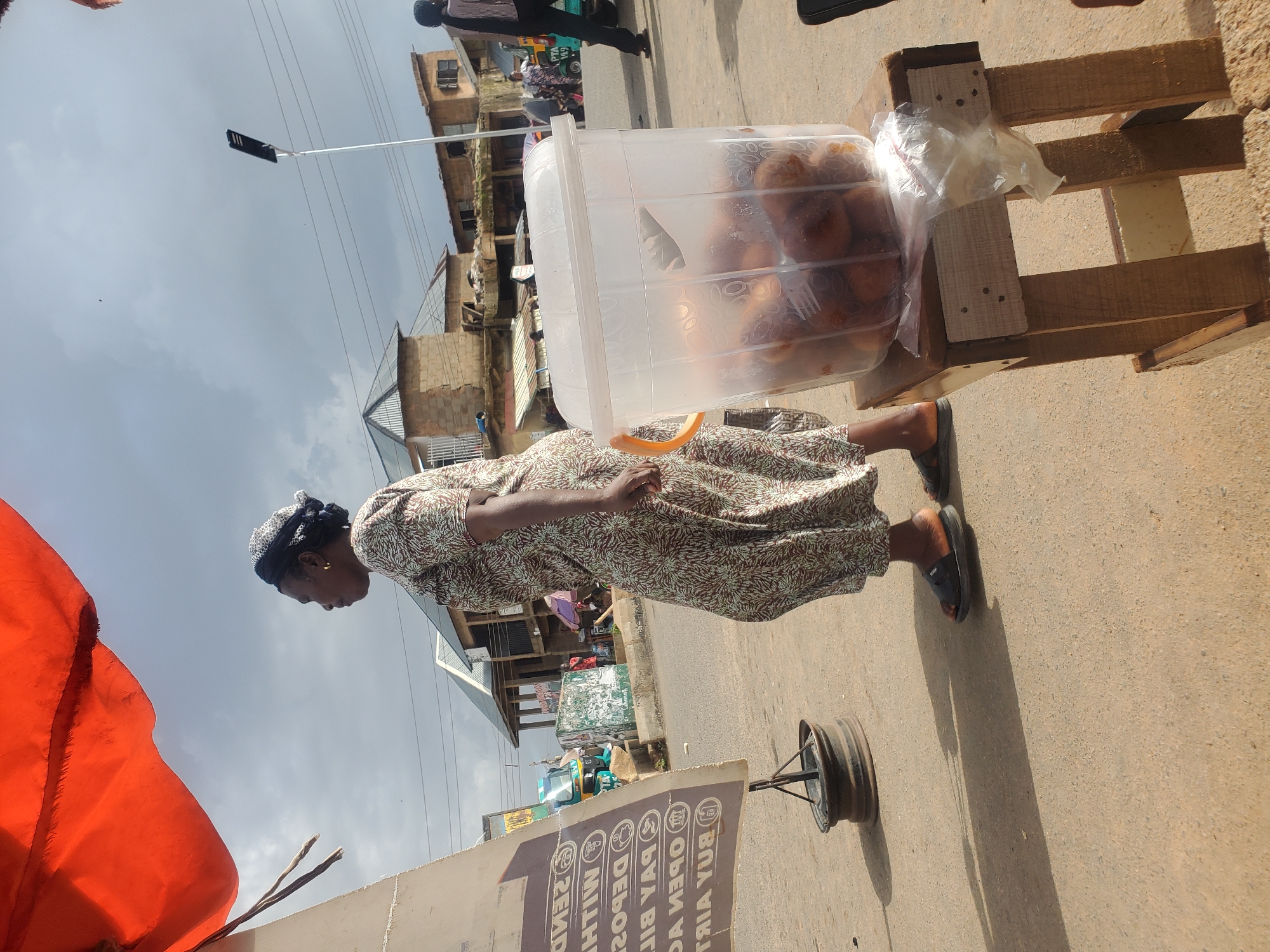 A woman stands beside a food stall in Gwong, Nigeria. The woman is wearing a patterned dress and a headscarf. She is looking down at a large plastic container filled with what appear to be fried dough balls, commonly known as puff puff. The container is placed on a wooden stand. In the background, there are buildings and a street with some traffic, including a tricycle. The sky is overcast, suggesting it might be a cloudy day. The time of day is not clearly discernible but appears to be daytime. A sign is visible in the foreground, but the text is partially obscured and difficult to read completely, though some words like "BUY AIR", "OPEN A", and "PAY BILL" are partially legible, suggesting it might be for a service provider. The overall scene depicts a street vendor in a market or roadside setting, engaged in selling food.