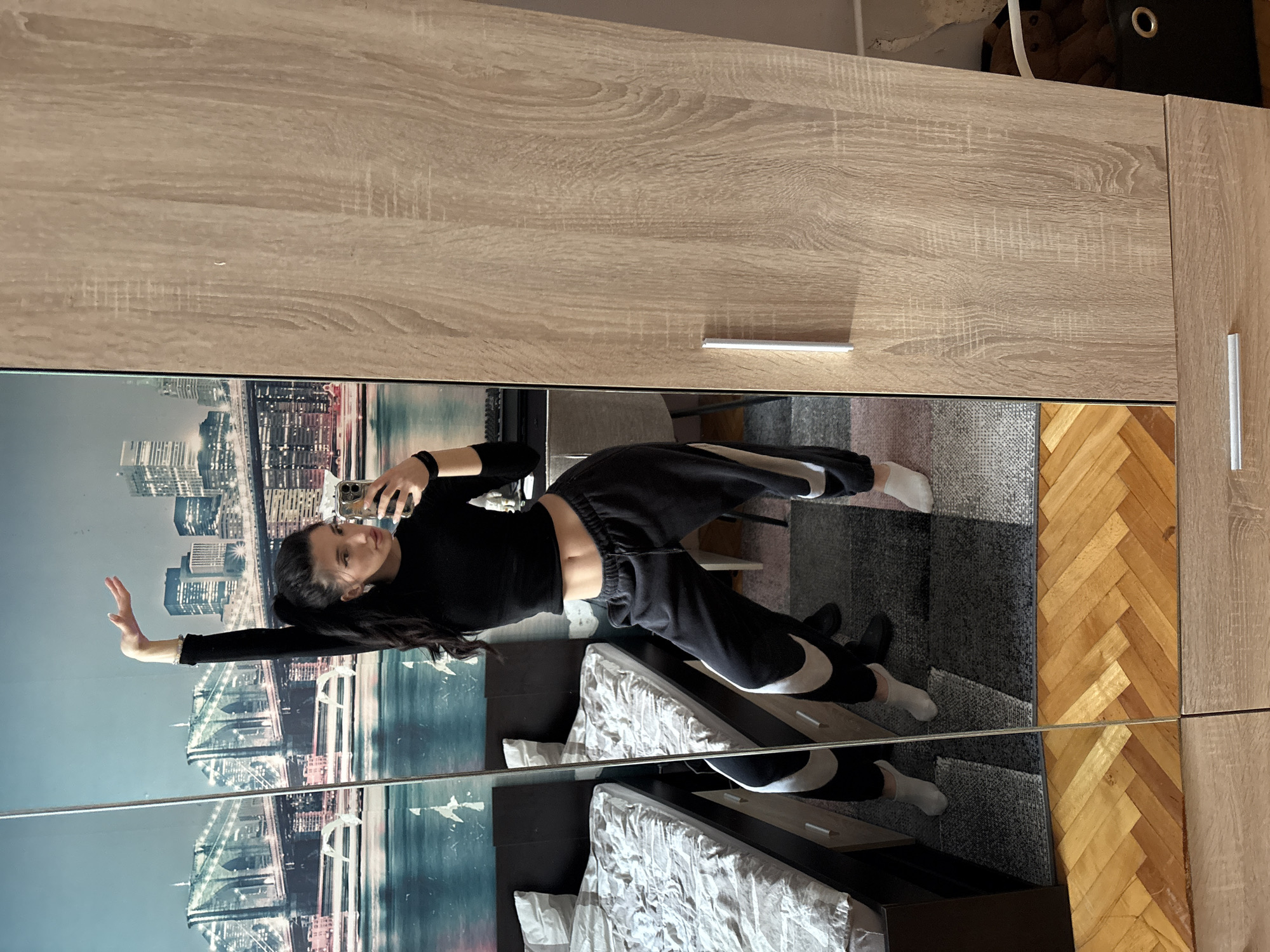 A woman is reflected in a large, mirrored wardrobe. She is wearing a black long-sleeved shirt and black sweatpants with a white Nike logo, and white socks. She is holding a smartphone, presumably taking a selfie. The wardrobe has a light wood finish and metal handles. The reflection shows a bed with white bedding, a dark headboard, and a gray rug. The floor is a herringbone pattern wood. The reflected background shows a cityscape and an image of the Tower Bridge in London. While the origin of the image is Sofia, Bulgaria, the city shown in the reflection is London.