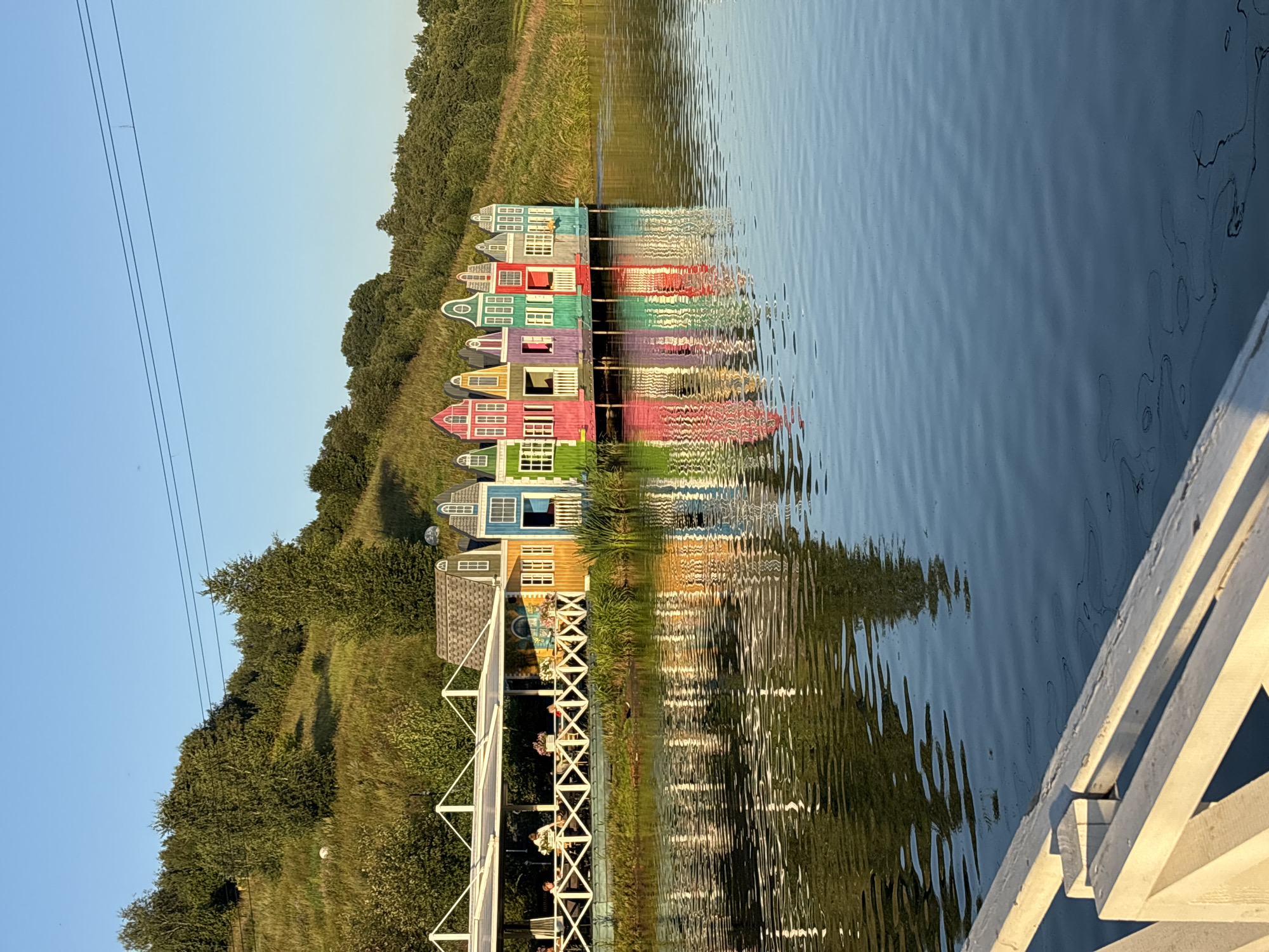 In Romashkovo, Russia, a picturesque scene unfolds by a calm body of water. The foreground features a white wooden railing, partially visible and leading the eye toward the main subject. Across the water, a row of brightly colored, miniature houses, reminiscent of Dutch architecture, stands on a pier. These whimsical structures, painted in vibrant hues of pink, purple, green, blue, yellow, and orange, are reflected in the water, creating a mirror image. Behind the colorful houses, a white-covered outdoor seating area with tables and chairs is visible, with a few people seated and enjoying the ambiance. The backdrop consists of a gently sloping, grassy hill covered in lush green trees, under a clear, bright blue sky. The sun casts a warm glow on the scene, indicating it is likely late afternoon or early evening.