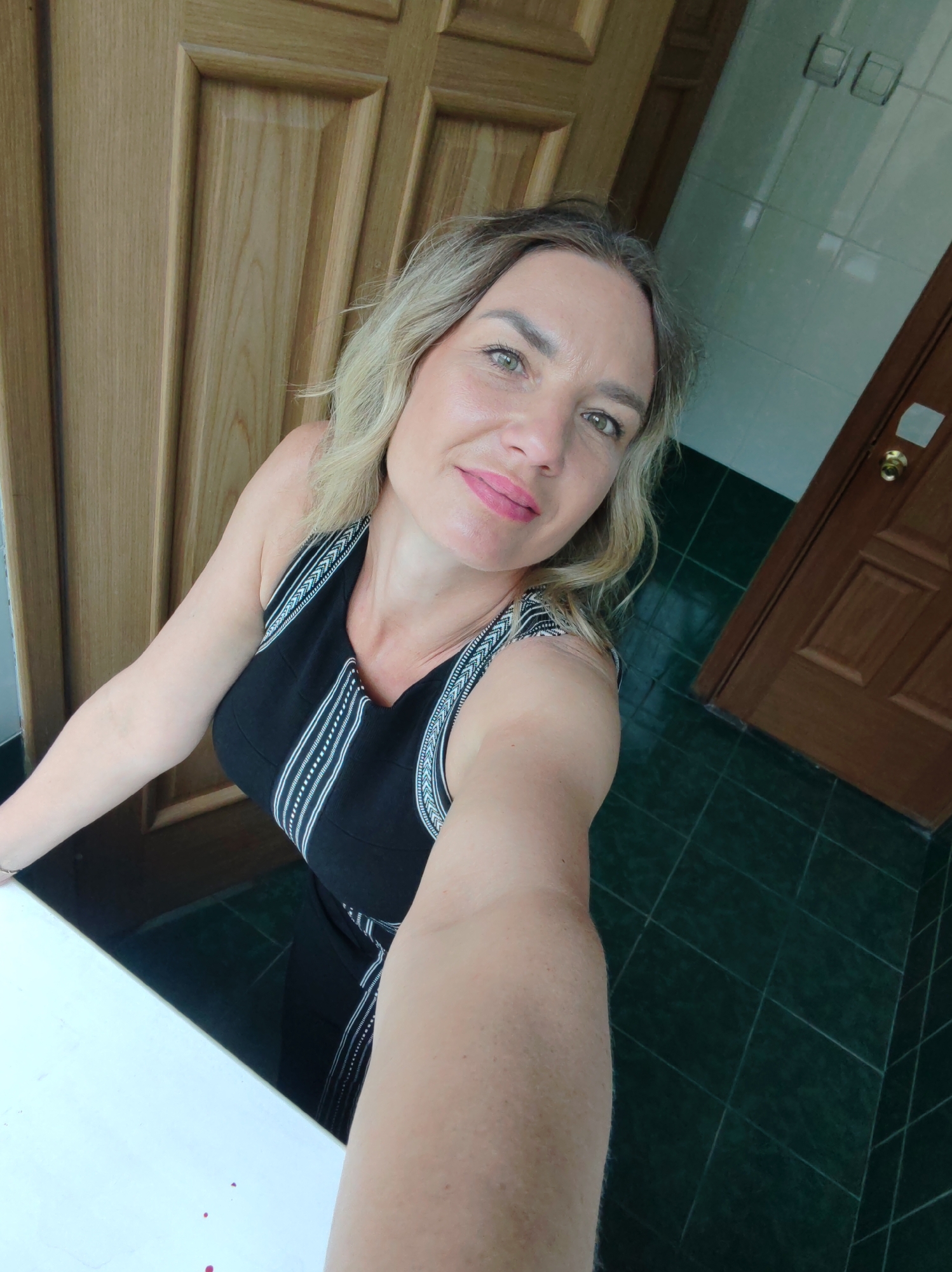 A fair-skinned woman with blonde hair is taking a selfie indoors. She is wearing a black dress with white vertical stripes on the front and shoulders. Behind her is a light-colored wooden door and a wall tiled with dark green tiles. A second wooden door is visible in the background on the right. The location in the city of Unknown, Unknown is unknown.