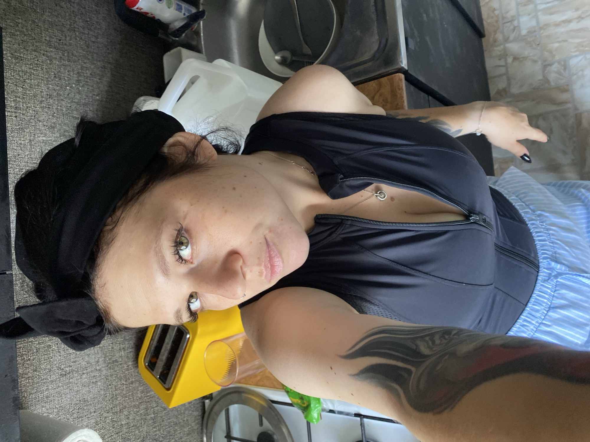 A woman with brown hair, wearing a black headscarf, a black zipped vest, and blue and white striped shorts, poses for a selfie in a kitchen. She has a tattoo on her left arm and is wearing a necklace with a circular pendant. In the background, a yellow toaster, a glass, a roll of paper towels, a sink with dishes, and large plastic containers are visible. The scene suggests a domestic environment.