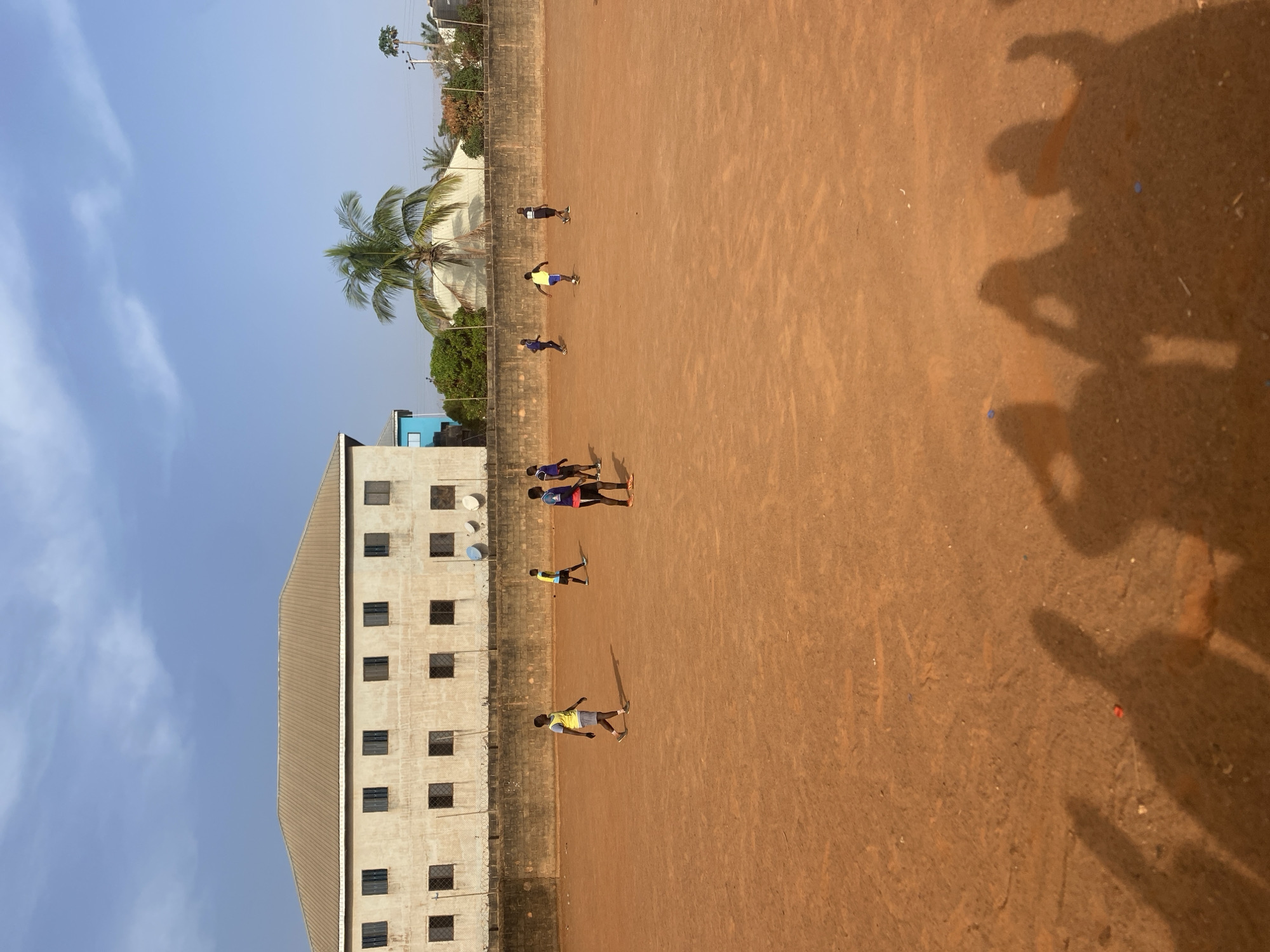 The image captures an outdoor scene in Jos, Nigeria, featuring a reddish-brown dirt field adjacent to a multi-story building under a clear blue sky. The field, likely a casual sports ground, is populated by several individuals, appearing to be children or young adults, dressed in casual sportswear like t-shirts and shorts. They are scattered across the field, engaged in an activity that suggests a game or exercise, possibly football, though no ball is visible.
To the left of the field, a tall, light-colored concrete building with numerous square windows rises, separated from the field by a low concrete block wall. This wall runs along the edge of the field, and behind it, various trees, including a prominent palm tree, are visible. In the far background, utility poles and wires can be discerned. The distinctive long shadows cast by the individuals on the field and the photographer in the foreground indicate either early morning or late afternoon sunlight. The sky is bright and clear, with only a few wispy clouds. The scene conveys a sense of everyday outdoor life and recreation. No discernible text is visible within the image.