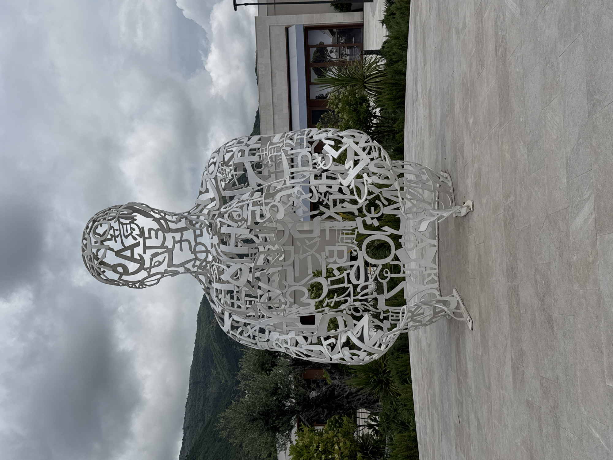 The image depicts a white metal sculpture of a seated figure composed of letters and symbols, likely located in Tivat, Montenegro. The sculpture is on a paved surface with a building and greenery in the background. A mountain is visible in the distance under an overcast sky.