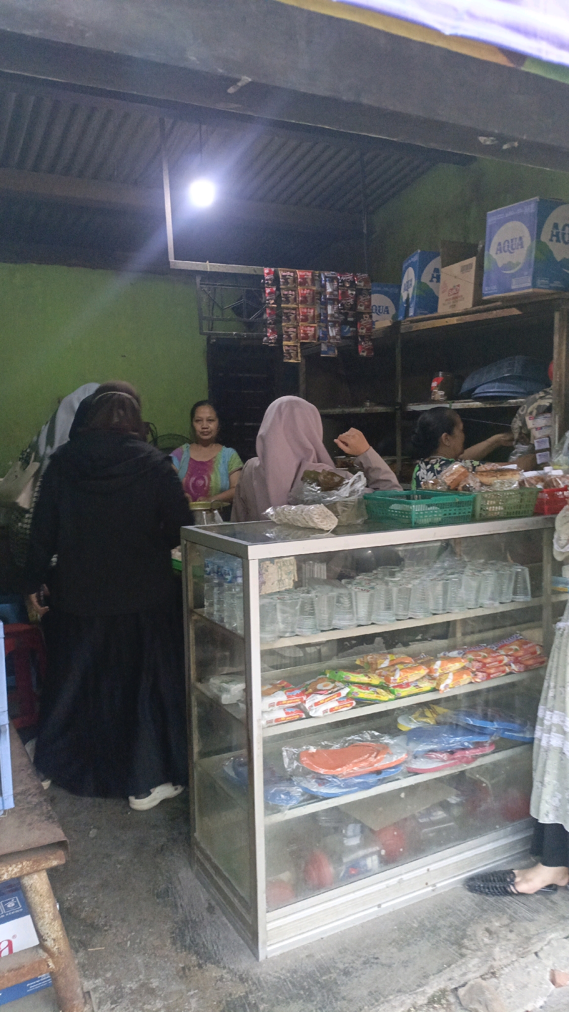 The image depicts an indoor scene at what appears to be a small shop or warung in Salatiga, Indonesia. A glass display case is prominent in the foreground, filled with rows of plastic cups and various packaged goods, including what look like instant noodles and possibly sandals on lower shelves. Behind the display case, several people are visible.
On the left, a person wearing a black hooded jacket and long dark skirt stands with their back mostly to the camera, facing towards a woman in a colorful short-sleeved top who is also behind the counter. To the right of this woman, another individual is wearing a light pink hijab and a brown long-sleeved top, with their right hand resting on the display case. Further to the right, another woman in a patterned shirt is partially visible, reaching towards a shelf.
The background shows shelves stocked with items, including boxes of "Aqua" bottled water, and rows of small packaged goods hanging from a rack. The environment appears somewhat cluttered, with a corrugated metal ceiling visible overhead and a green wall on the left. The lighting suggests it might be daytime, with a bright light fixture hanging from the ceiling. The floor is concrete and appears worn.