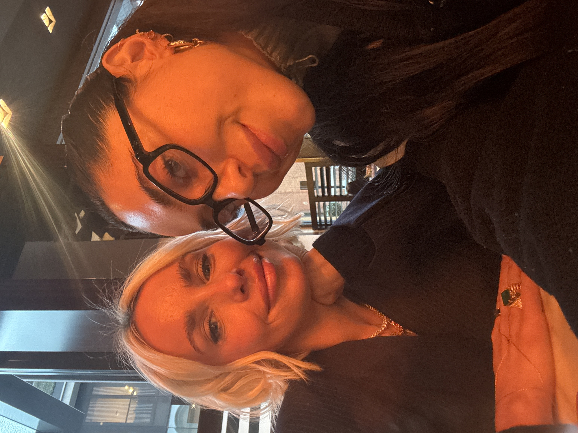 The image presents a close-up selfie of two adult females indoors, in what appears to be a high-end establishment within the city of London, United Kingdom. The exact venue or address is not discernible from the visual evidence.
The individual positioned on the left has fair skin, short, wavy blonde hair, and is wearing a dark pinstripe blazer over a lighter garment. She accessorizes with two layered necklaces, one featuring small clear stones and another with small red stones, and a prominent gold ring with a large green stone on her right ring finger. Her right hand rests on a light-colored surface, propping her chin. She displays a faint smile and looks directly towards the camera.
The individual on the right has darker skin, dark hair pulled back from her face, and is wearing black-framed eyeglasses. She is dressed in a dark turtleneck garment, visible zipper pull at the collar. Her left ear is adorned with multiple gold hoop and stud earrings. She also exhibits a faint smile, looking towards the camera, and is positioned closer to the lens, suggesting she is operating the camera device.
The environment is characterized by warm, artificial lighting, casting an orange hue across the subjects. The background on the left reveals a large window overlooking a modern building exterior with vertical architectural elements, glass panels, and a balcony featuring greenery, consistent with contemporary urban architecture found in London. The background on the right is darker, showing subtle hints of horizontal slats or blinds and a dark vertical structural element within the interior. Square, recessed light fixtures emitting a warm glow are visible on the ceiling.