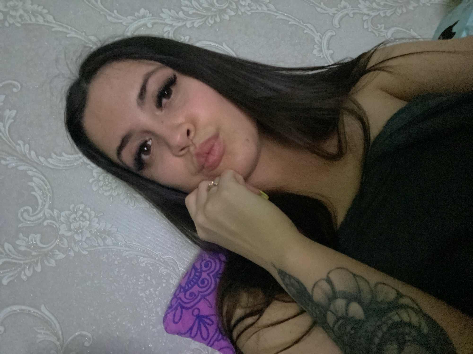 A young adult woman is lying down in a room in what could possibly be the city of Chaikovsky, Russia. She is lying on a pillow with a purple patterned design, and is partly covered by a dark-colored garment. Her face is close to the camera, and she has dark hair, eyelashes that appear to be enhanced, and is pursing her lips. Her right hand is near her face, and a ring is visible on one of her fingers. Her left arm is visible, with a tattoo covering a portion of it. The wall behind her features a subtle floral pattern.