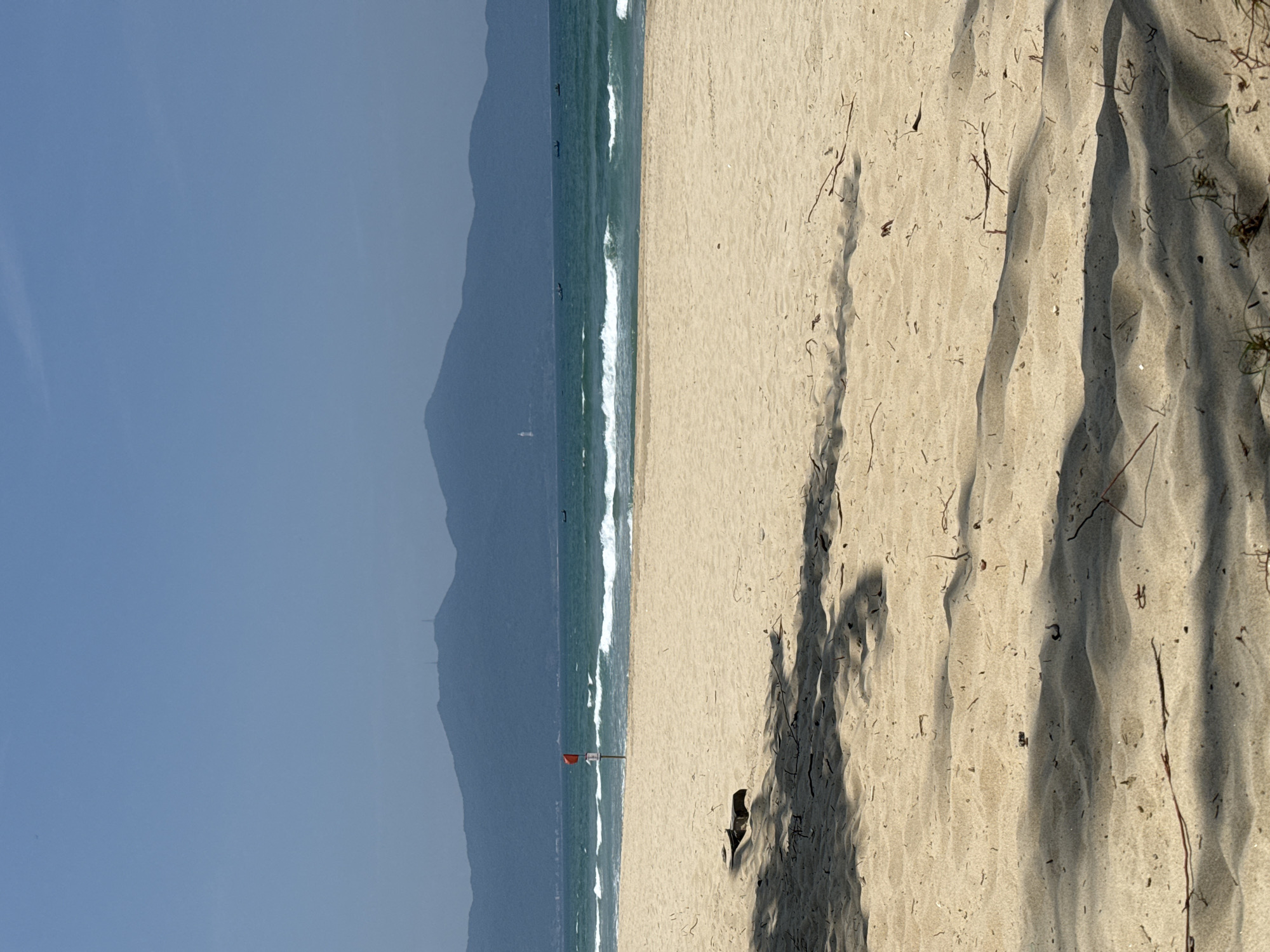 A horizontal photograph depicts a beach scene, possibly in Đà Nẵng, Vietnam. The left side of the image shows the sea, with waves and a red flag on a pole near the shore. In the far background are hills or mountains under a hazy sky. The right side shows sand, with patterns of light and shadow. There are no people visible in the image. Vegetation and scattered twigs lie on the sand.