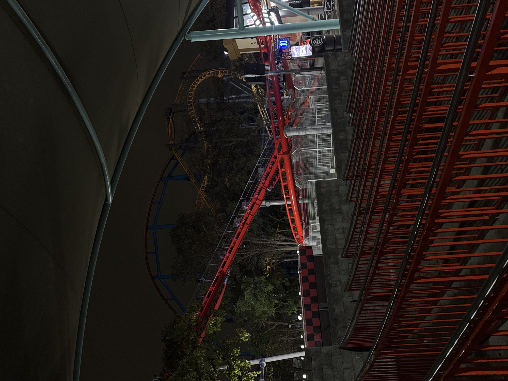 At night in Mexico City, Mexico, a view of roller coasters. The red roller coaster structure is prominently visible, along with portions of other roller coasters with blue and yellow tracks in the background. To the left, a curved, dark structure. In the foreground, a red ladder-like structure. Trees are visible within the composition.