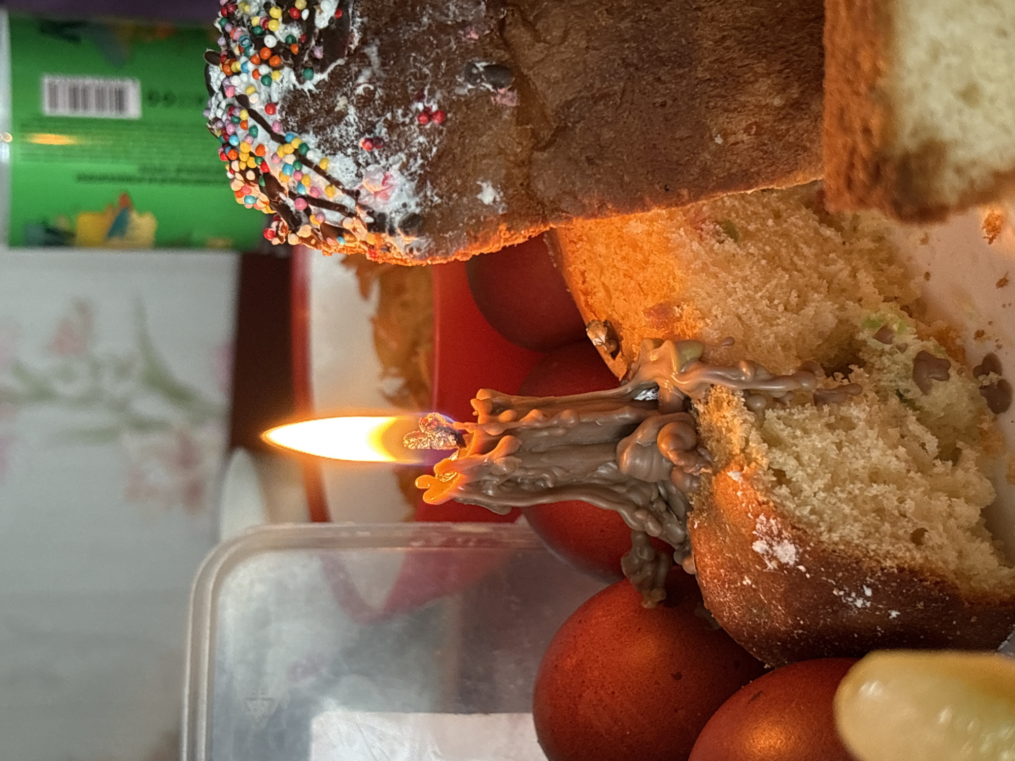 A close-up indoor shot shows food, likely bread or cake, in front of a lit candle with dripping wax, resting on what appears to be dyed eggs. The cake has sprinkles and chocolate and is partially cut, revealing the crumb. The candle is almost entirely melted, with the wax running down the cake. The background is blurry, with a container and part of a can visible. No people are visible in the frame. The location is not possible to determine from the image itself, though Пожарское сельское поселение is a potential location based on the request.