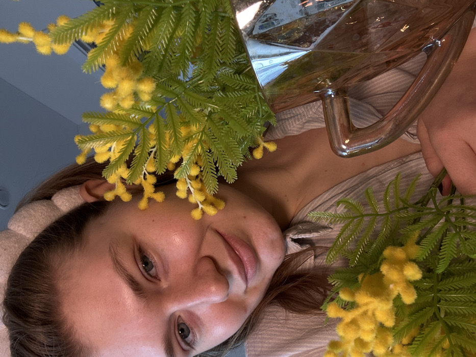 A woman's face, angled, is visible among green and yellow foliage. Her complexion is fair, with clear skin and visible eyebrows and eyelashes. She wears a light-colored headband. Her lips are slightly parted in a smile. Above her, a transparent glass object with a metallic handle reflects the surrounding environment. She is holding the flowers in her right hand. The scene appears to be an intimate, close-up perspective.