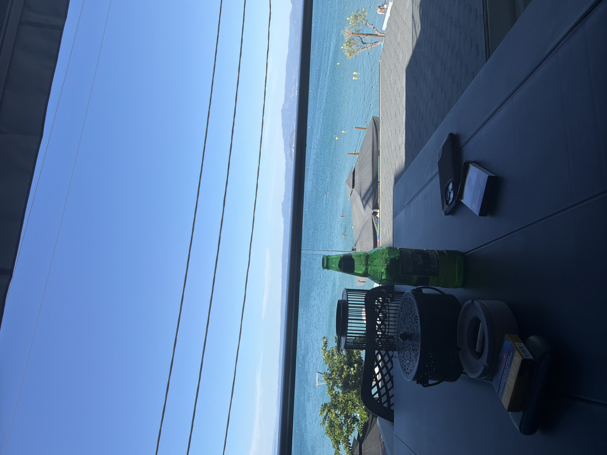 This image captures a scene from a balcony overlooking a body of water, possibly a lake or sea, under a clear, bright blue sky. The viewpoint is framed by a window with vertical blinds. Visible across the water is a shoreline with mountain ranges in the distance. The water has a turquoise hue and is dotted with yellow buoys and possibly other watercraft.
On the balcony, a dark-colored table occupies the foreground. It holds various items including a green glass beer bottle, a metal container with a decorative lid, a round ashtray, and a pack of cigarettes. A phone and a card are next to each other on the table as well. A patterned metal chair can be seen behind the items.
The precise location within Pallini Municipal Unit, Greece, cannot be definitively determined from the image alone. However, the presence of a lake or sea view from a balcony suggests a coastal or lakeside setting, which could be typical of certain areas within the municipality.